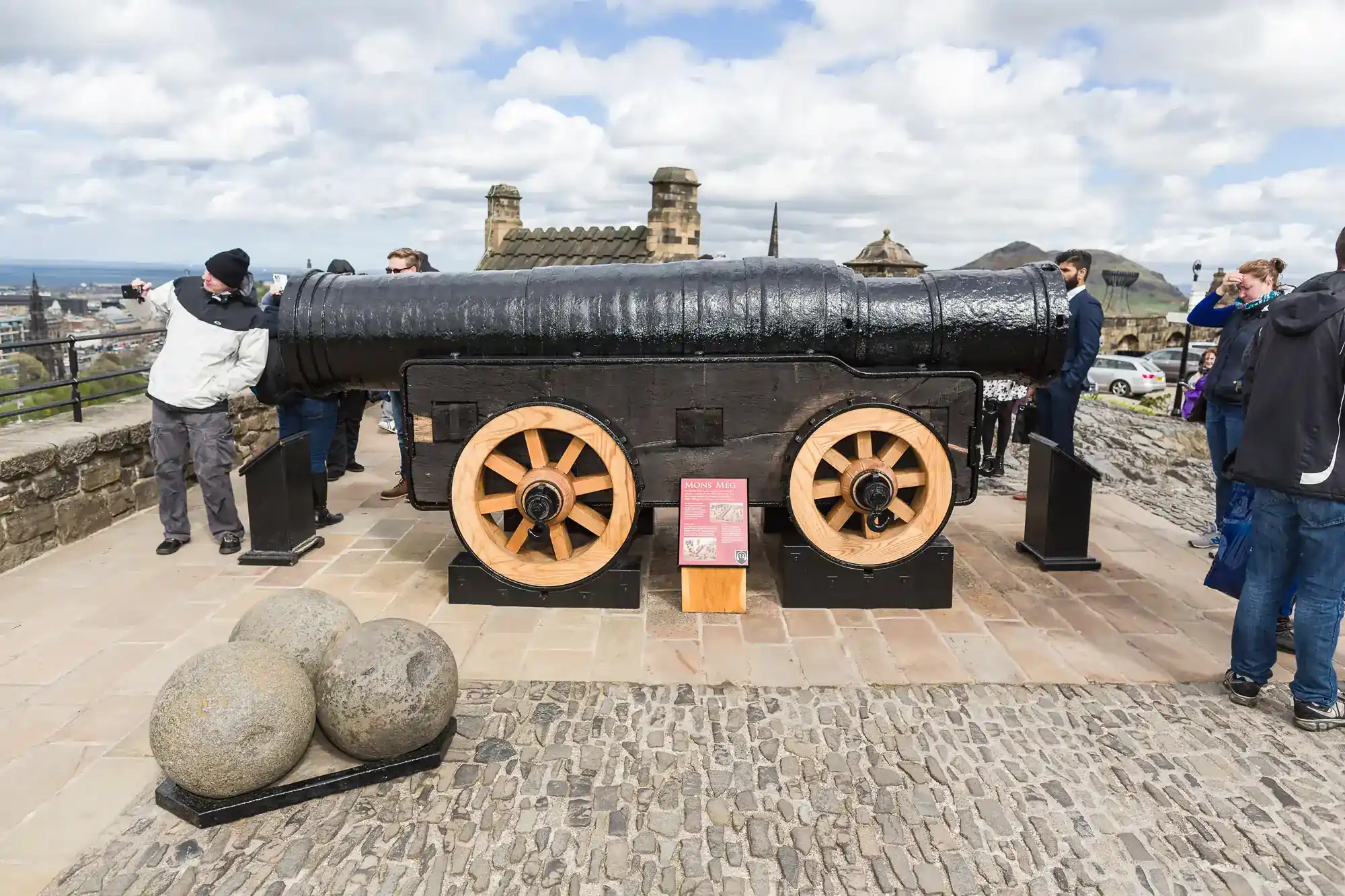 A large black historic cannon with wooden wheels is displayed on a stone platform outdoors. Several people stand nearby, and three large cannonballs are beside it. Cloudy sky and distant buildings are visible in the background. A red informational sign stands by the cannon.