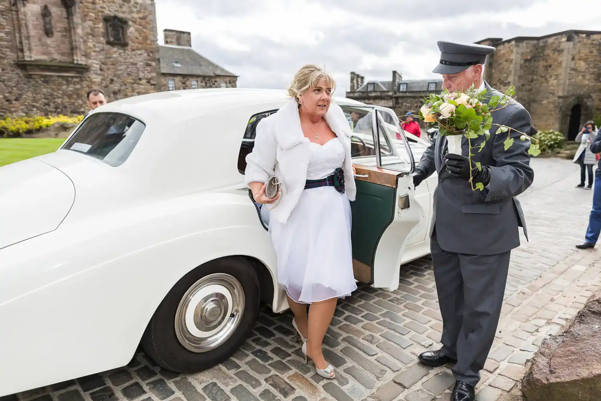 A woman in a white dress and shawl steps out of a vintage white car onto cobblestones, assisted by a chauffeur in a grey uniform holding flowers. The setting is outside an old stone building on a cloudy day, with people and greenery in the background.