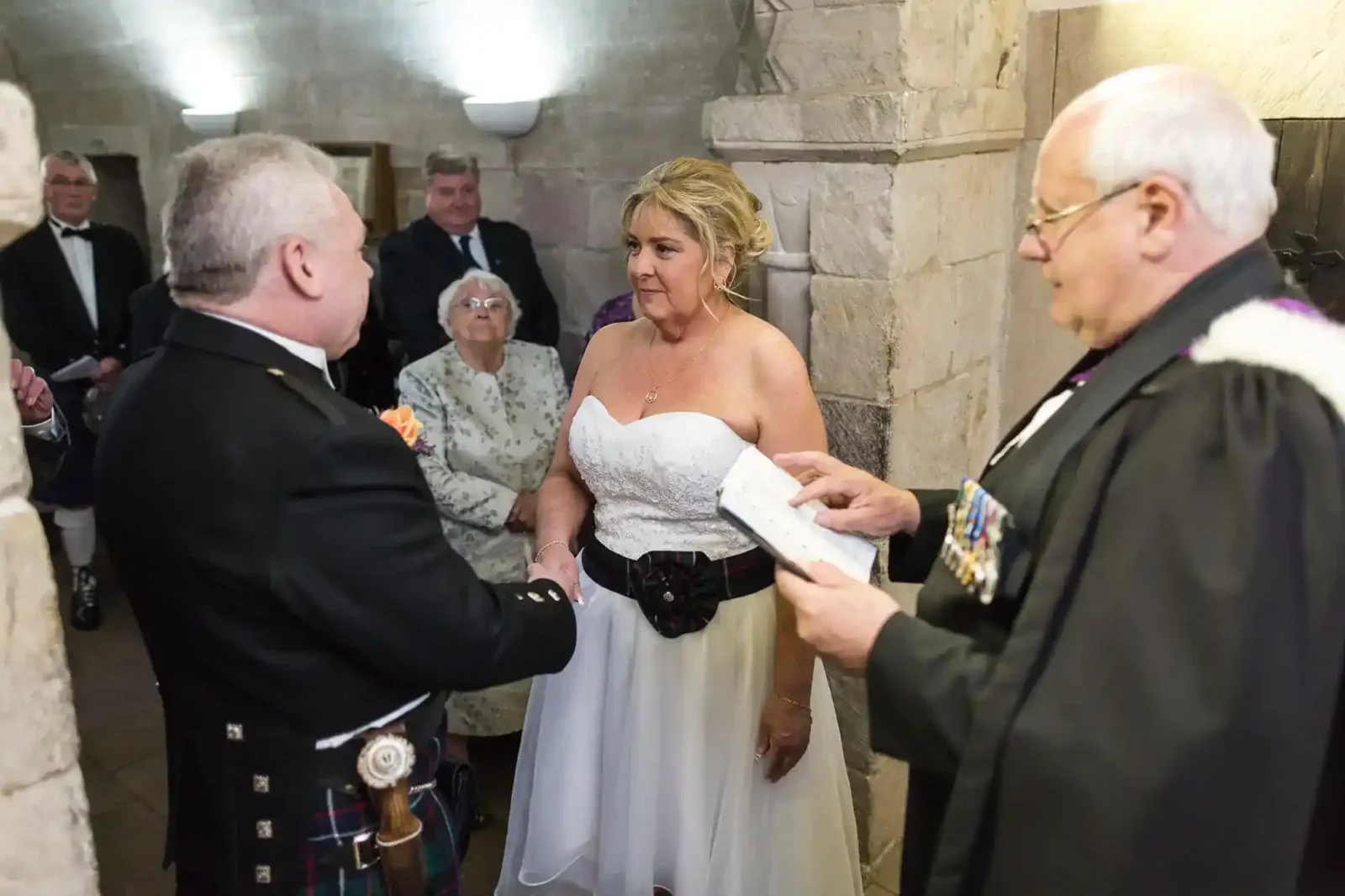 Bride in a white dress and groom in Scottish attire stand facing each other, holding hands during a wedding ceremony. An officiant reads from a book. Onlookers, including an elderly woman, watch in a stone-walled room at St Margaret’s Chapel at Edinburgh Castle..