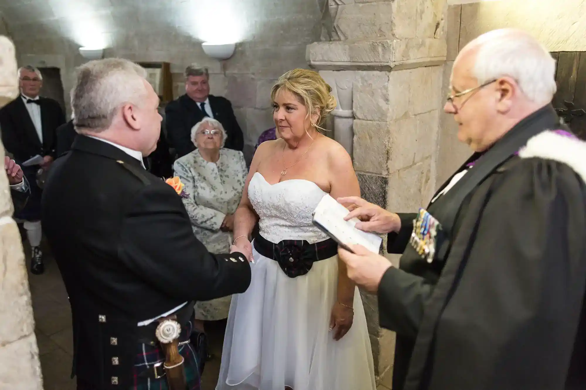 Bride in a white dress and groom in Scottish attire stand facing each other, holding hands during a wedding ceremony. An officiant reads from a book. Onlookers, including an elderly woman, watch in a stone-walled room.