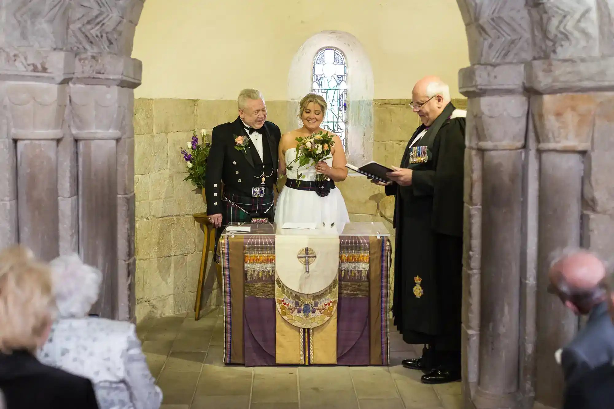 A bride holds a bouquet, standing between an older man in a kilt and a priest reading from a book. They are at an altar in a stone chapel, with ornate pillars framing the scene and a stained glass window behind them. Seated guests watch the ceremony.
