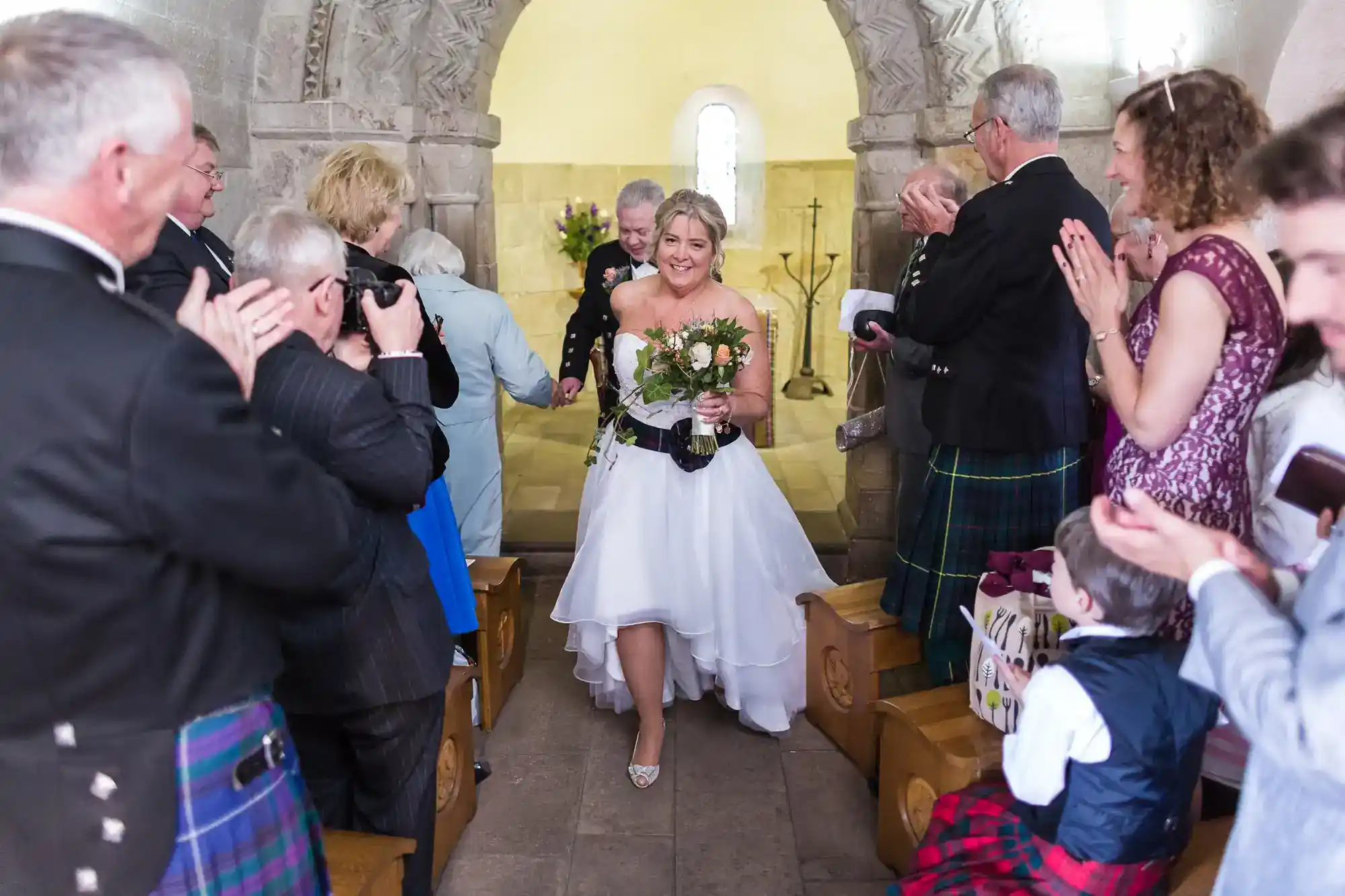 A smiling bride in a white dress walks down a stone church aisle, holding a bouquet. Guests wearing formal and traditional attire clap, smile, and take photos. Light streams in from a window at the front of the church.