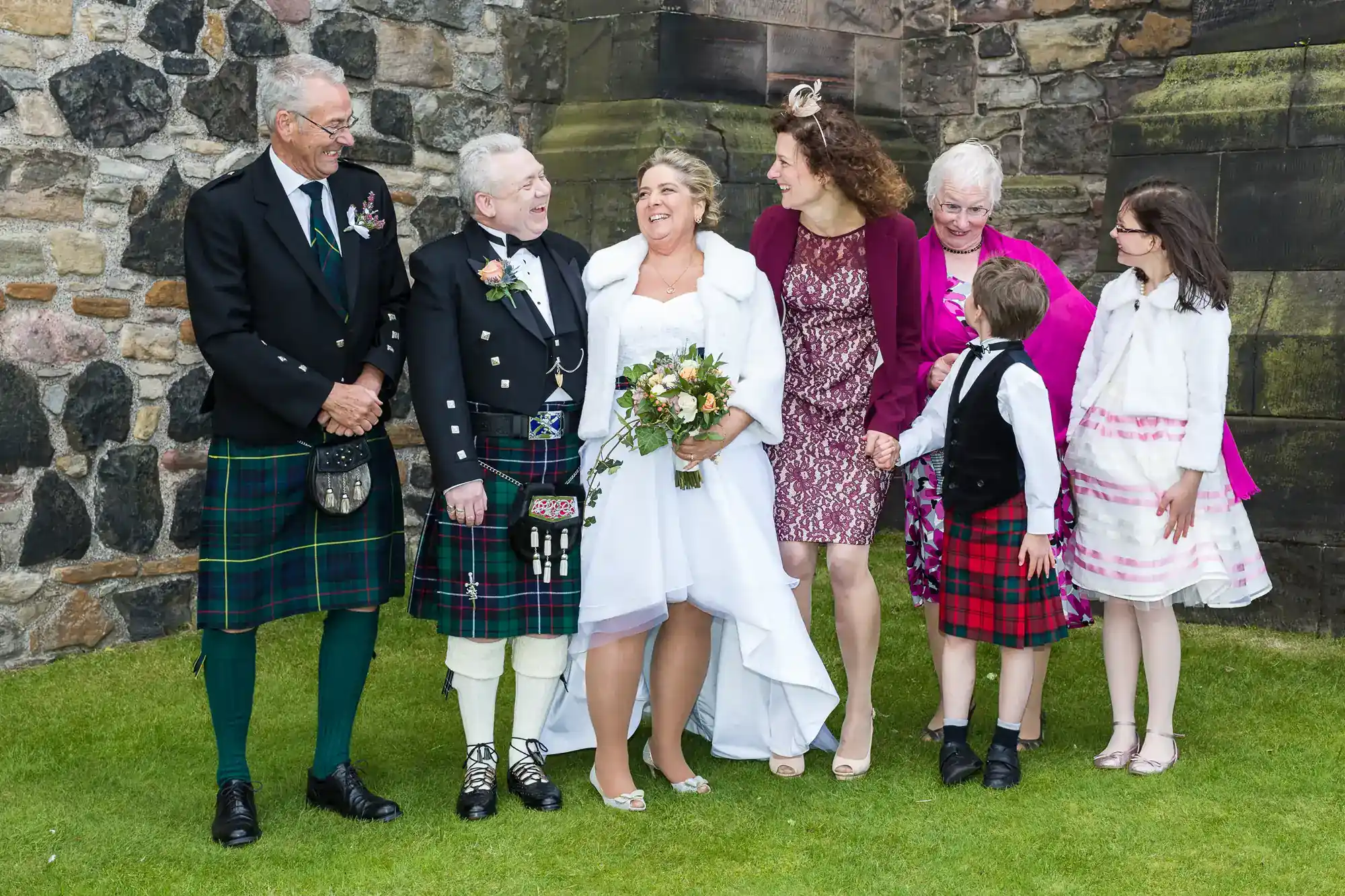 Six people pose happily outdoors at a wedding; two men in kilts, a bride in a white dress holding flowers, two women in colourful dresses and fascinators, a boy in a kilt, and a girl in a white dress. They stand on grass in front of a stone wall.