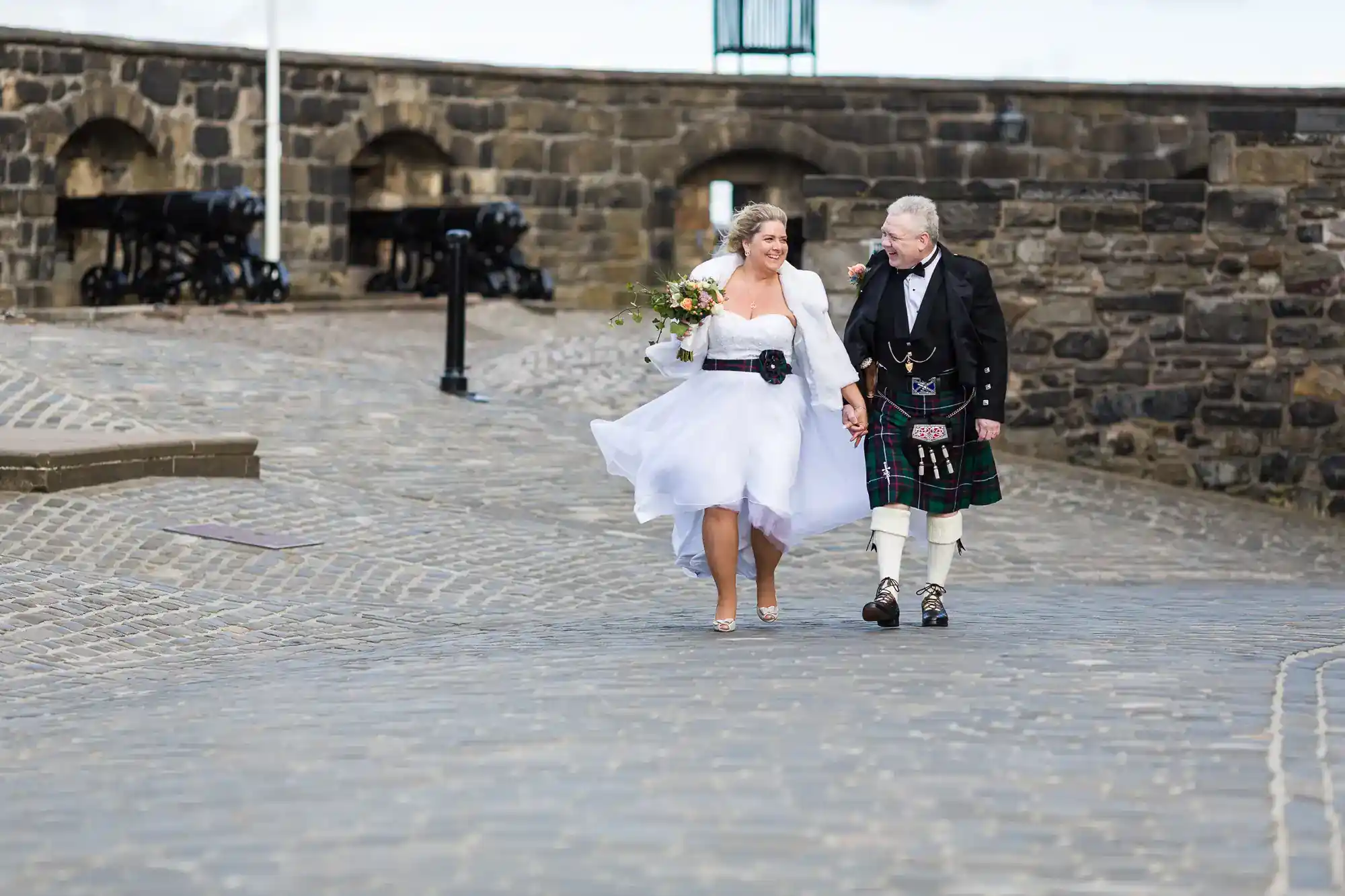 A bride in a white dress and a groom in a traditional Scottish kilt walk hand in hand on a cobbled path by an old stone wall, smiling at each other. The bride holds a bouquet, and both look joyful and relaxed.