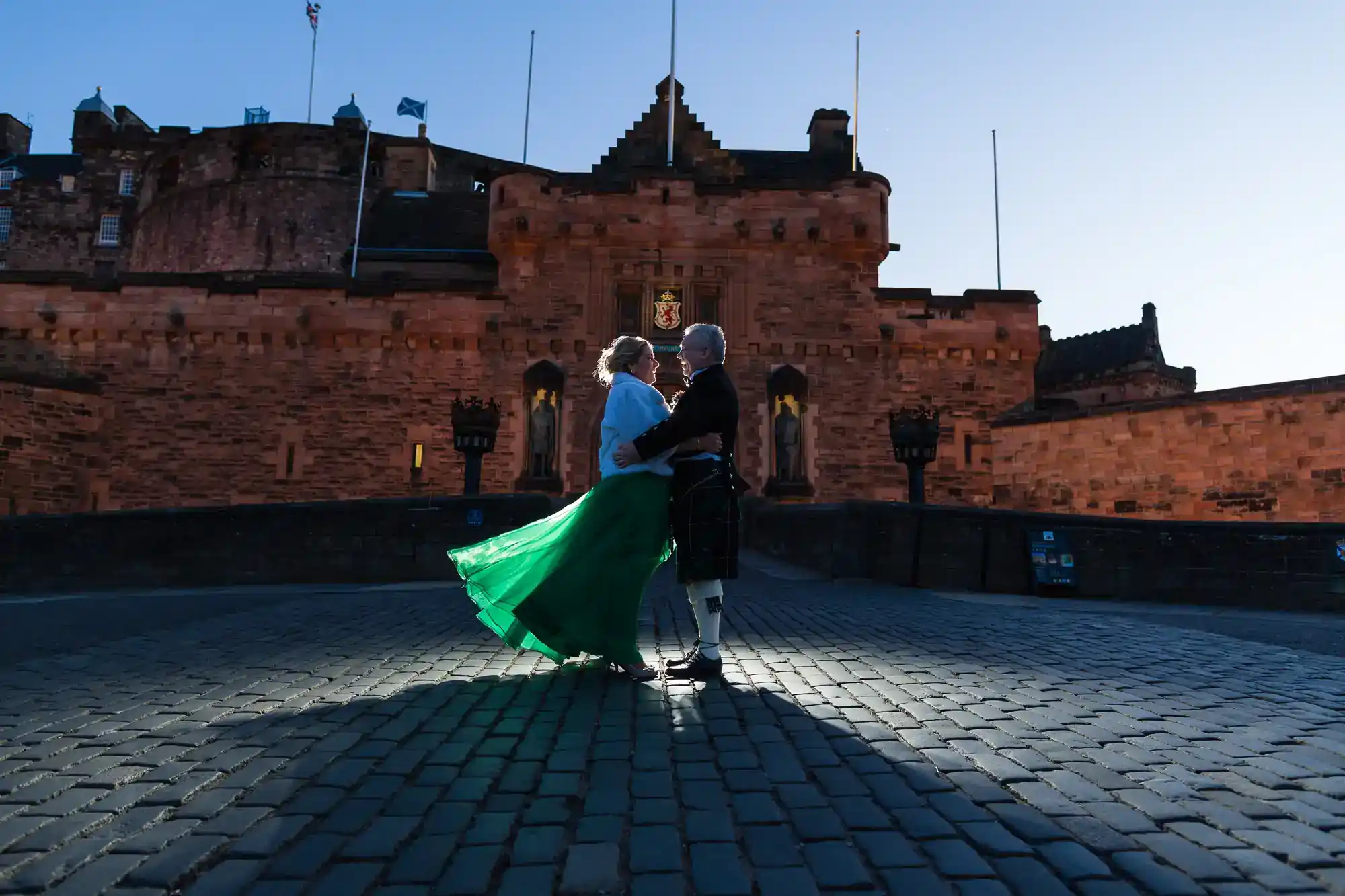 An older couple dance on cobblestones in front of a historic stone castle at dusk. The woman wears a long green skirt that flows outward, and the man wears a kilt. The castle’s flags are at half-mast, and soft evening light creates dramatic shadows.