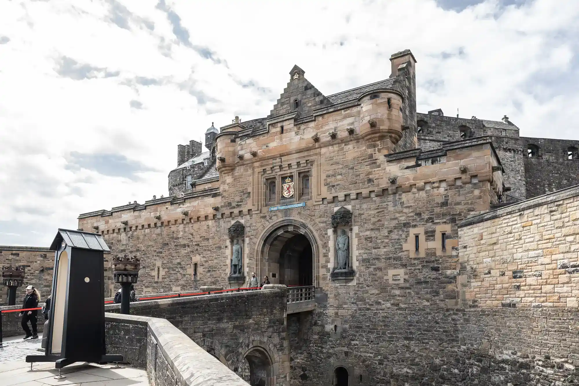 Stone fortress entrance with arched gateway, statues on either side, and a crest above. A stone bridge leads up to it, and a guard post sits to the left. The sky is partly cloudy. Several people are visible near the entrance.