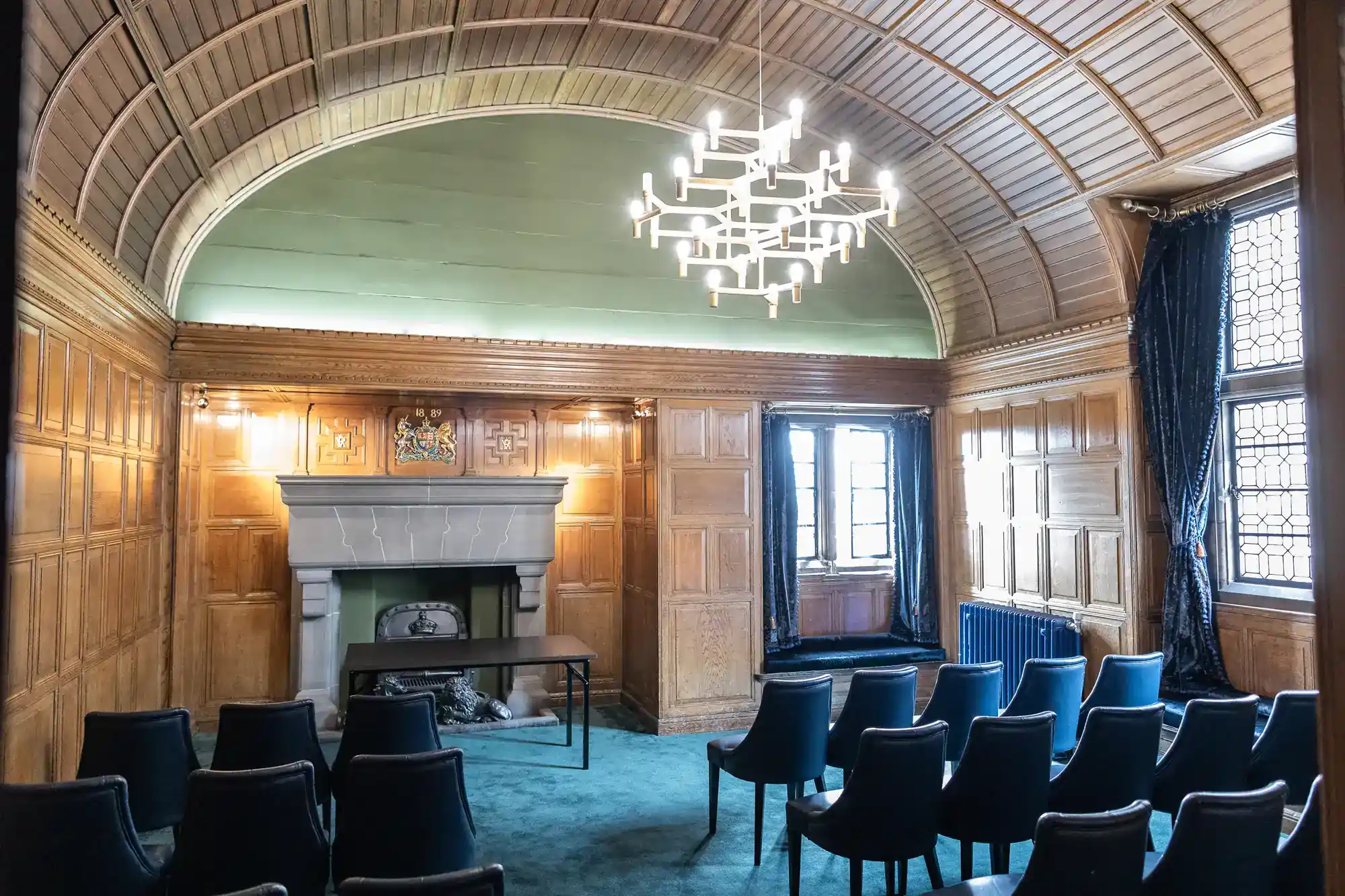 A wood-panelled room with arched ceiling, blue carpet, and rows of blue chairs facing a fireplace and table. A modern chandelier hangs above. Sunlight streams through tall windows with dark curtains.