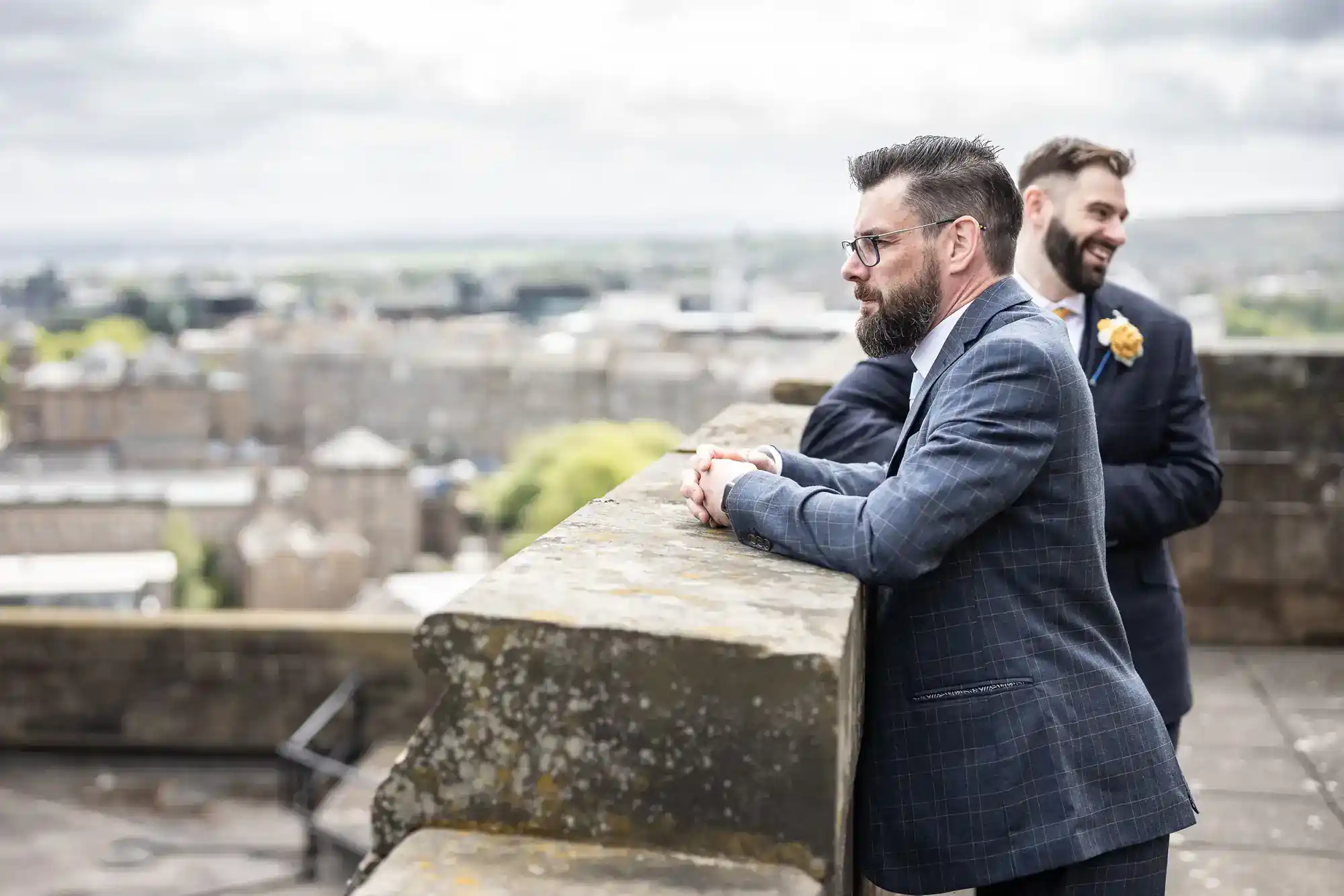 Two men in suits stand on a stone terrace overlooking a cityscape. The man in front leans on the railing, wearing glasses and a blue checked suit. The man behind him, smiling, has a yellow buttonhole. The background is blurred with cloudy skies.