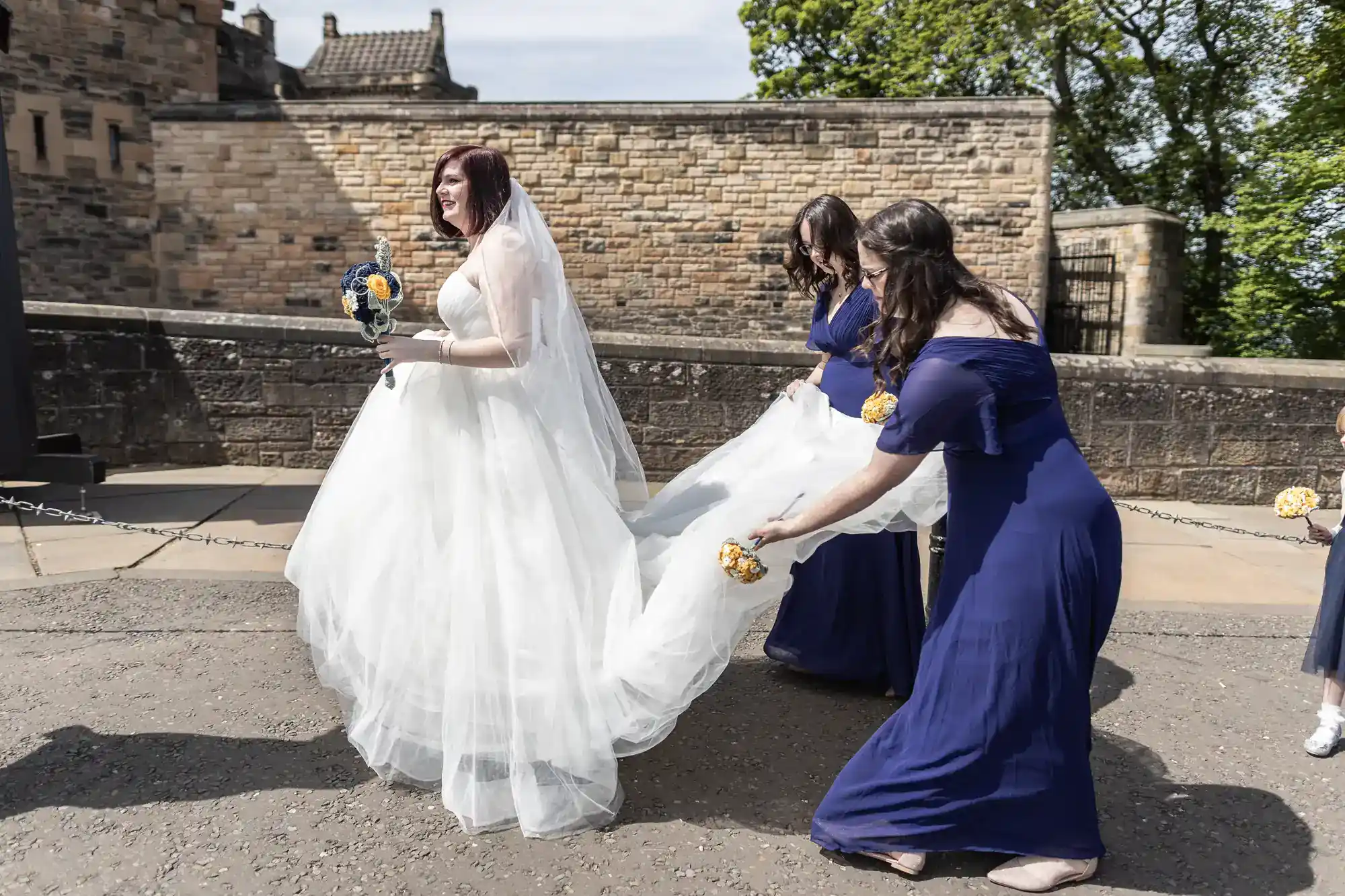 A bride in a white dress holds yellow flowers as two bridesmaids in blue dresses adjust her train outside on a sunny day, near a stone wall and green trees.