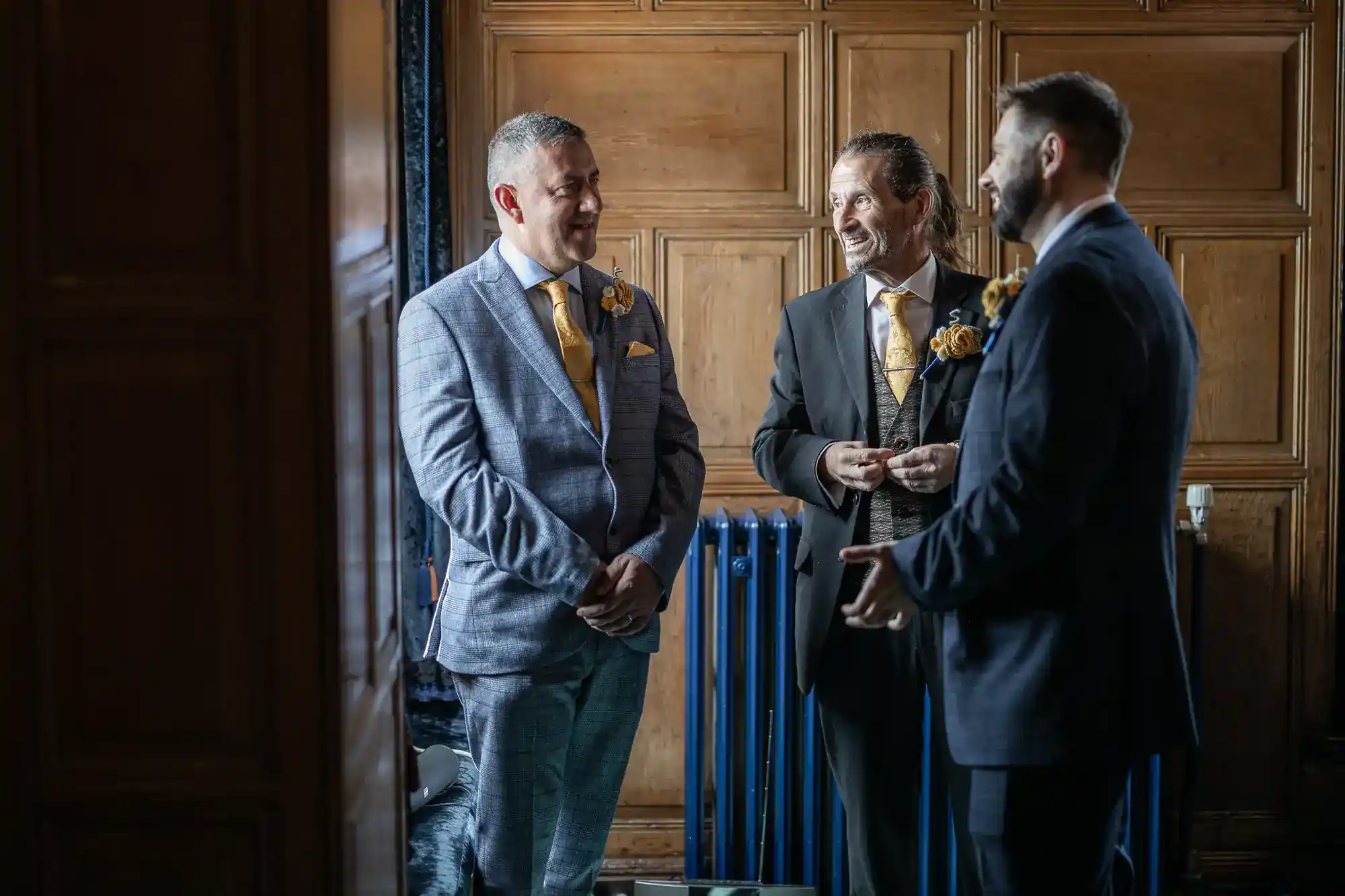 Three men dressed formally in suits and yellow ties stand indoors, smiling and chatting. They are in a wood-panelled room with blue radiators. Sunlight softly illuminates them from the left, creating a warm and friendly atmosphere.