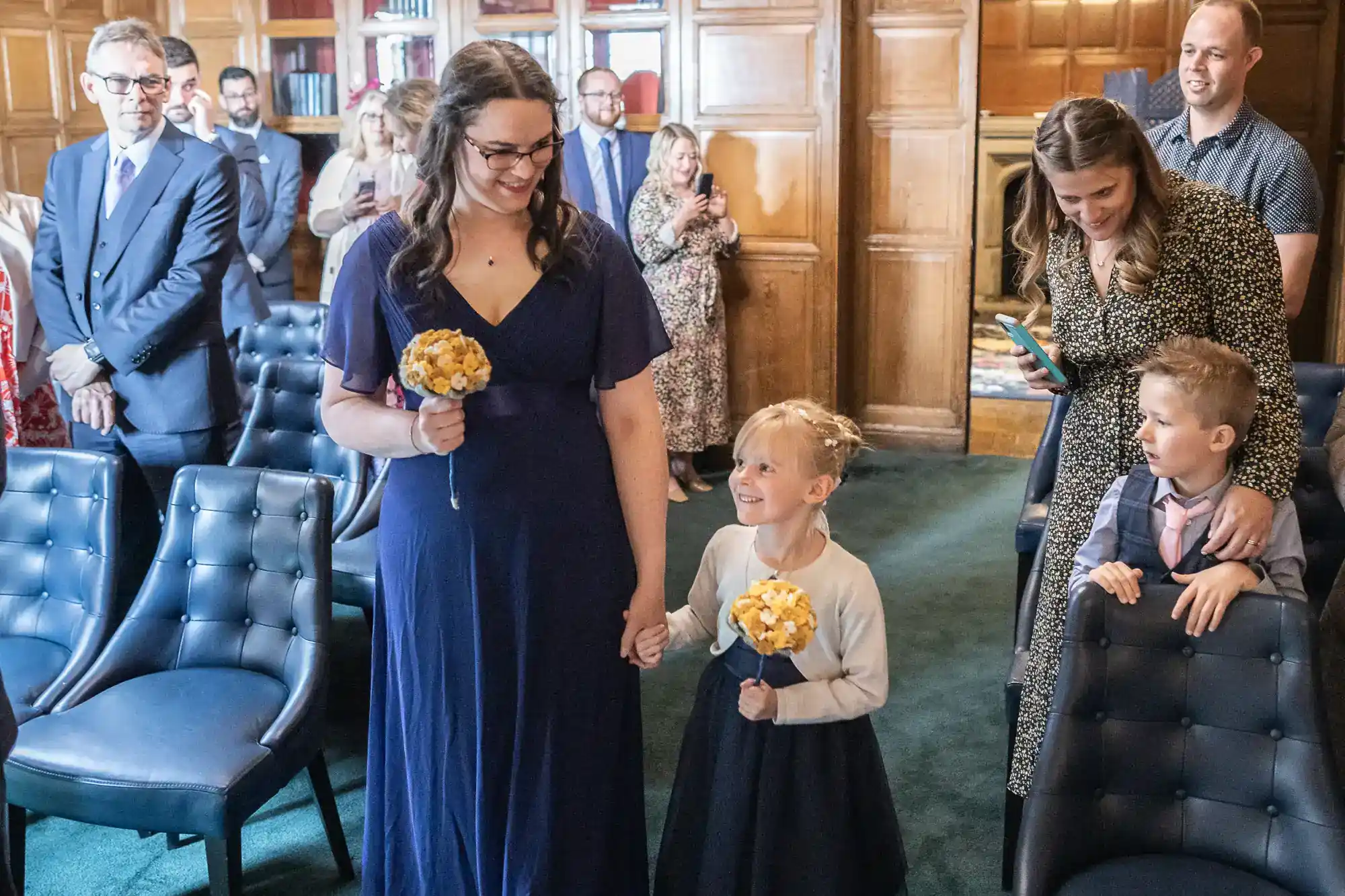 A woman in a navy dress and a child holding her hand walk down an aisle in a wood-panelled room, both holding yellow bouquets. People sit and stand nearby, smiling and looking on. Shelves of books and wood panelling are visible in the background.