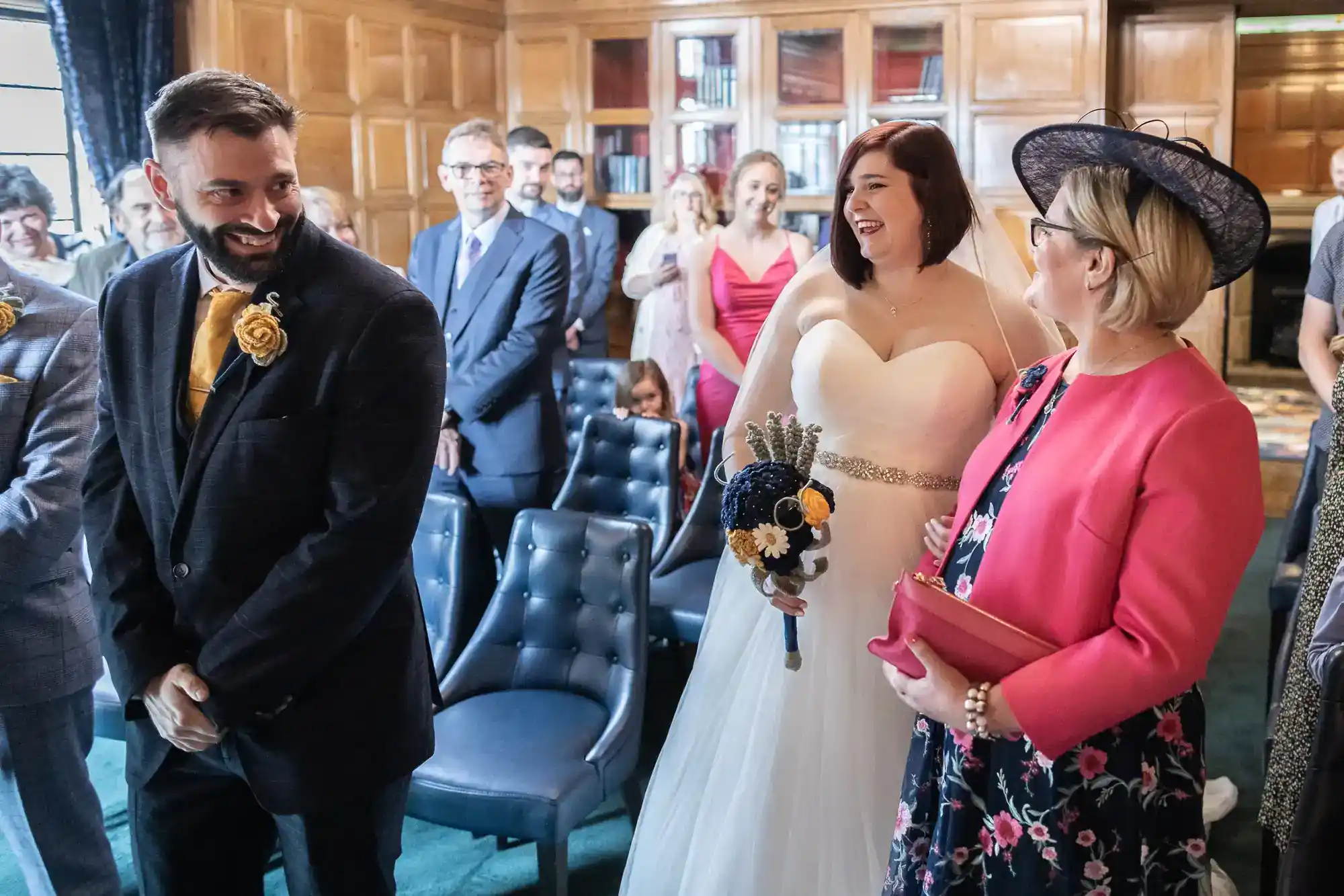 A bride in a white dress and groom in a checked suit smile at a woman in a pink jacket and hat during a wedding ceremony. Guests in formal attire sit and stand among wooden-panelled walls and bookshelves, creating a warm, joyful atmosphere.