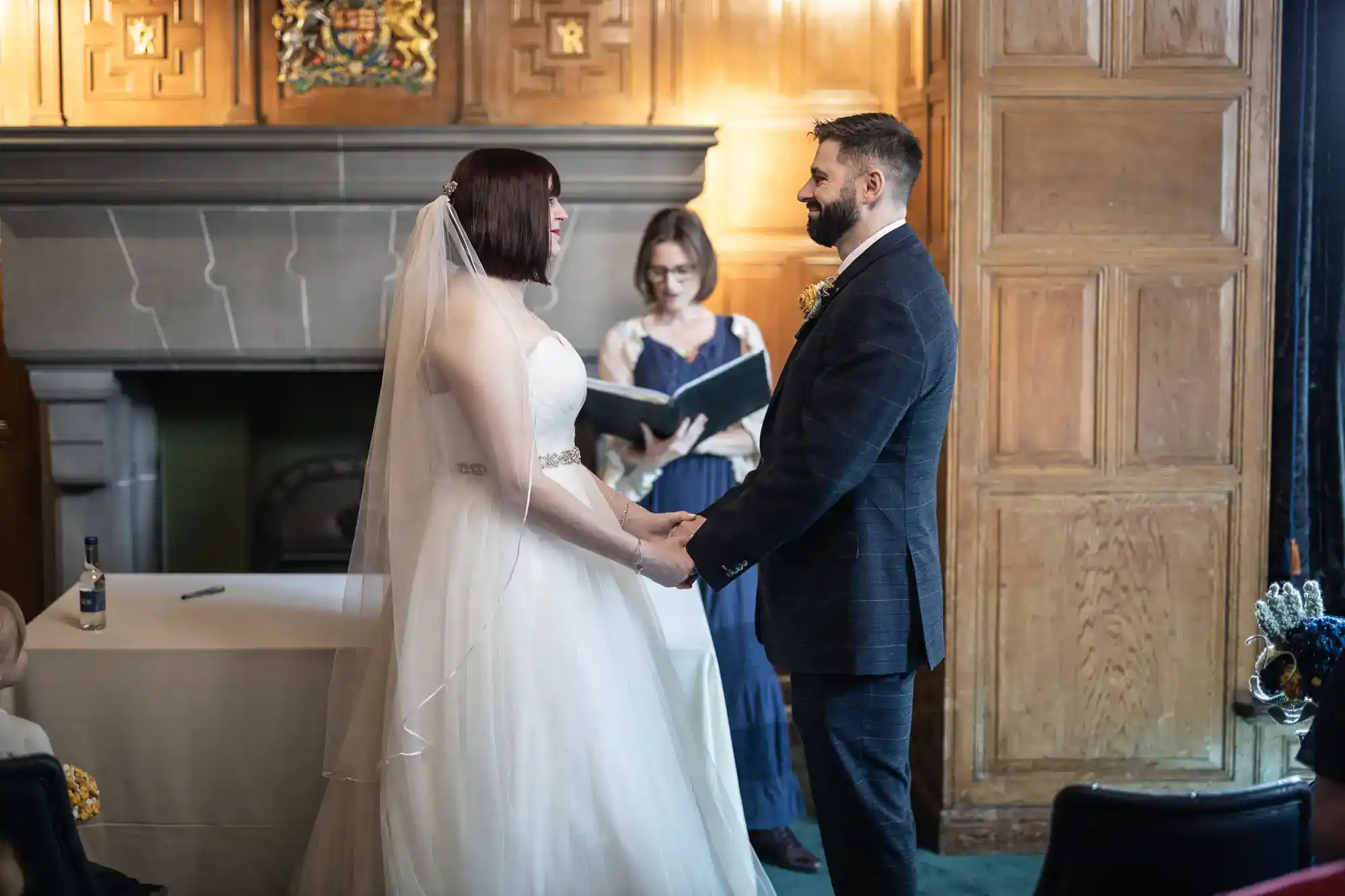 A bride in a white dress and veil faces a groom in a dark suit as they hold hands during a wedding ceremony. An officiant stands behind them with an open book. They are indoors, in front of a fireplace and wood-panelled wall, smiling at each other.