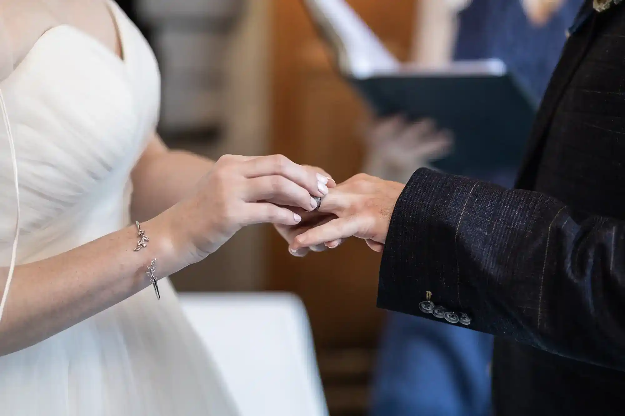 A bride in a white dress places a ring on her partner’s finger during a wedding ceremony. The groom wears a dark suit. Both hands are visible in the foreground, whilst a person holding a book or script is slightly blurred in the background.