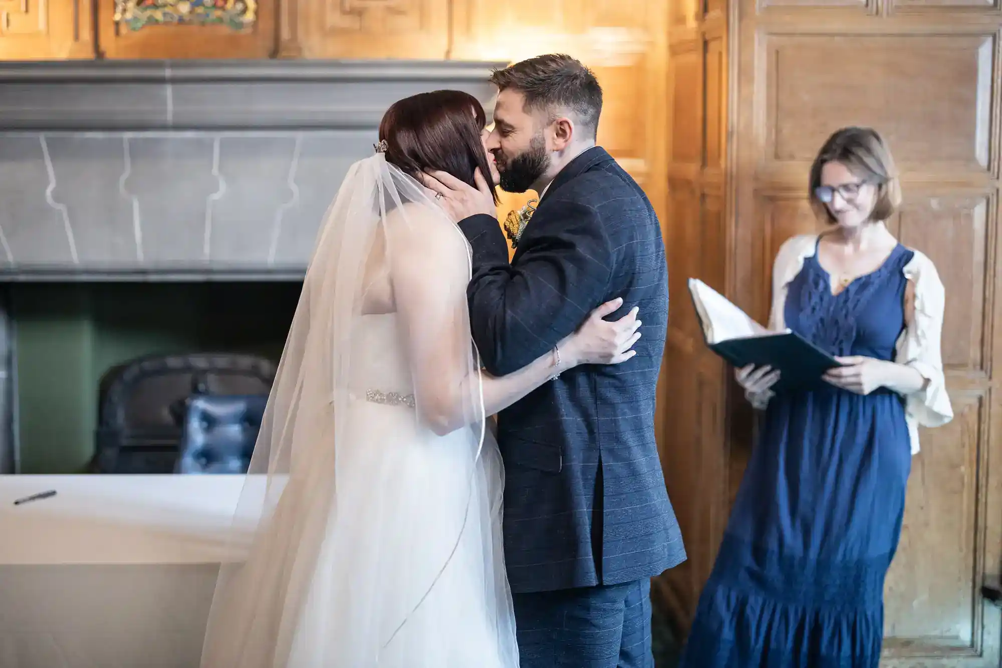 A bride in a white dress and veil kisses a groom in a blue suit. Both stand in a wood-panelled room. In the background, a woman in a blue dress holding an open folder smiles as she watches the couple share their moment.