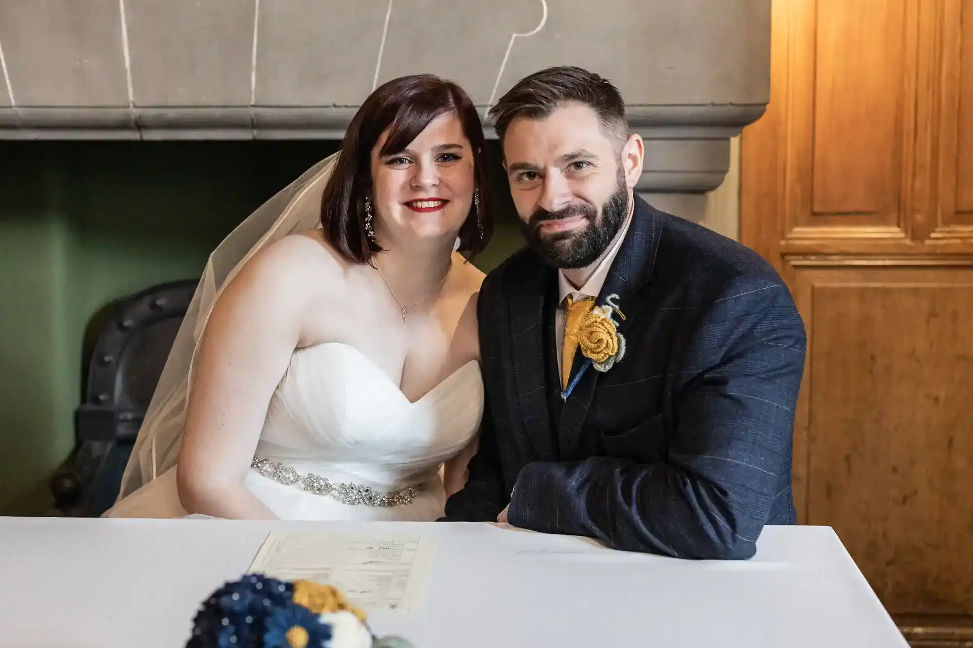 A bride in a white dress and veil sits beside a groom in a dark suit with a yellow buttonhole. They smile at the camera while seated at a white table, signing a document. Warm-toned wooden and stone walls are in the background. A bouquet is in the foreground.