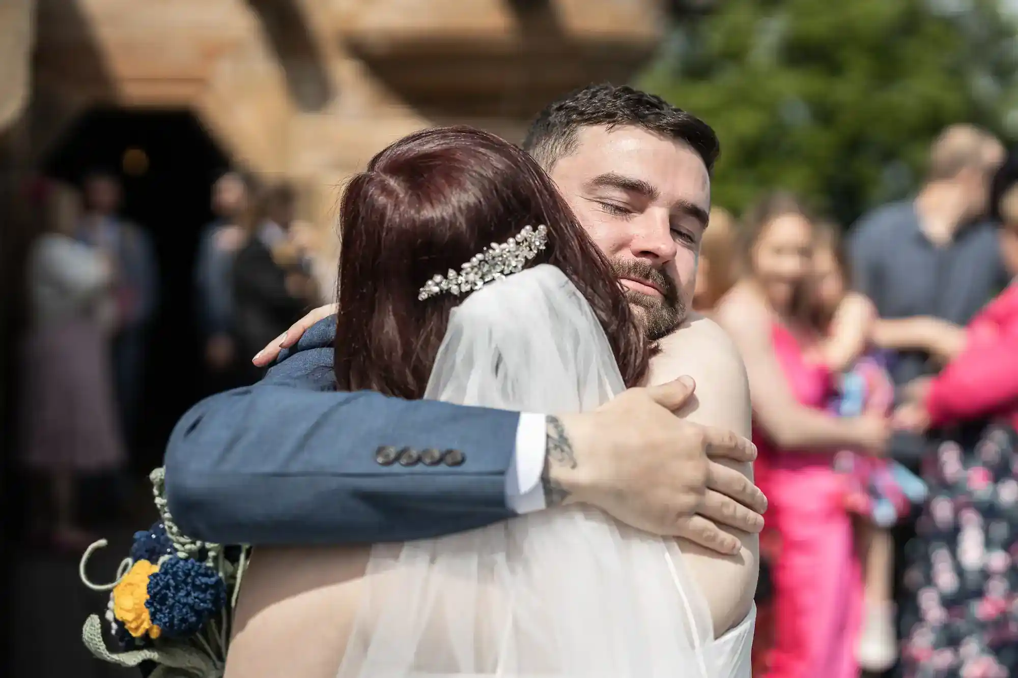 A bride with a jewelled hairpiece and veil hugs a man in a blue suit outside, both smiling. People in colourful outfits stand blurred in the background under sunlight. The scene conveys warmth and celebration at a wedding.