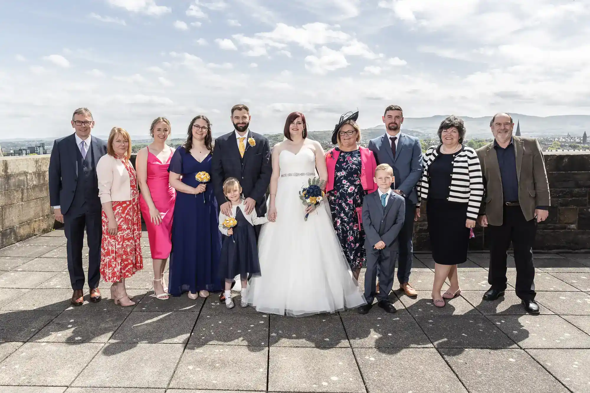 A wedding group poses outdoors on a sunny day. The bride in a white dress stands centre, flanked by the groom and guests in formal attire. Two children, a girl in blue and a boy in a suit, stand in front. Mountains and a blue sky fill the background.