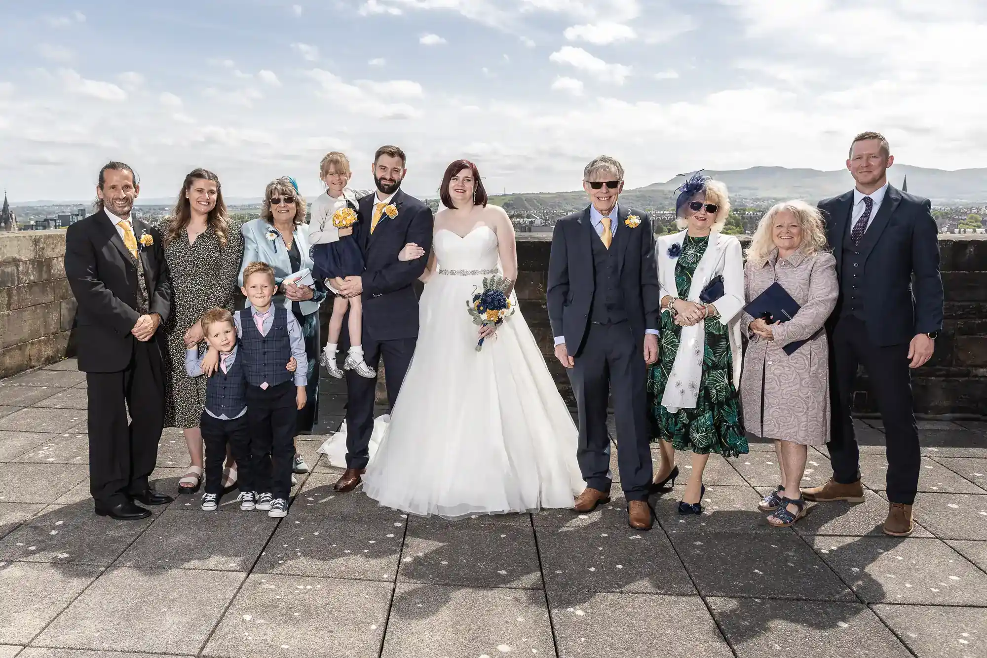 Twelve people stand on a rooftop, dressed in formal wedding attire. The bride wears a white dress and holds a bouquet; the groom stands beside her. Four children and others in colourful dresses and suits smile in daylight, with hills and sky in the background.
