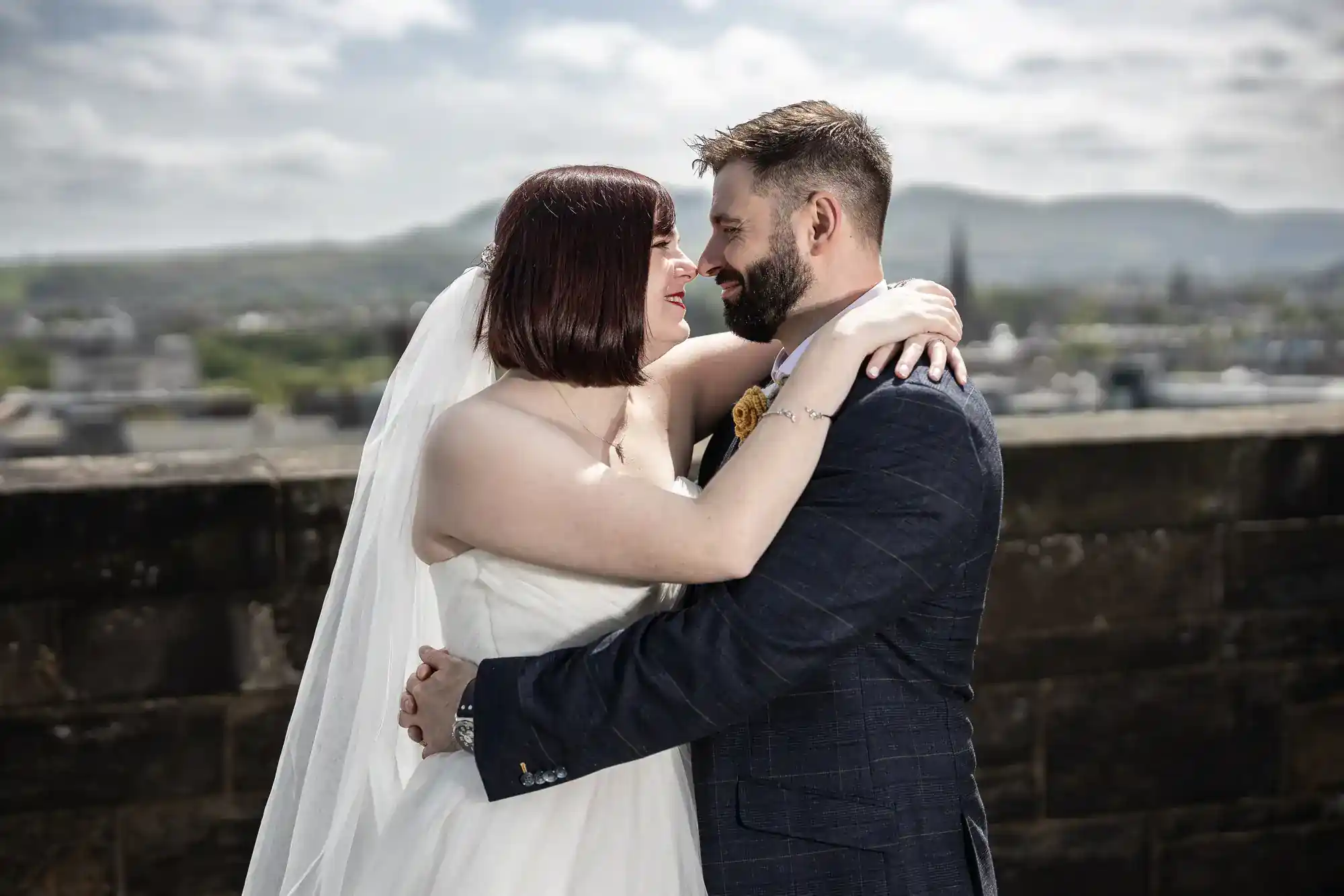 A bride and groom embrace and smile at each other outdoors, with the bride in a white dress and veil and the groom in a dark suit. A stone wall and blurred cityscape with hills are visible in the background under a cloudy sky.