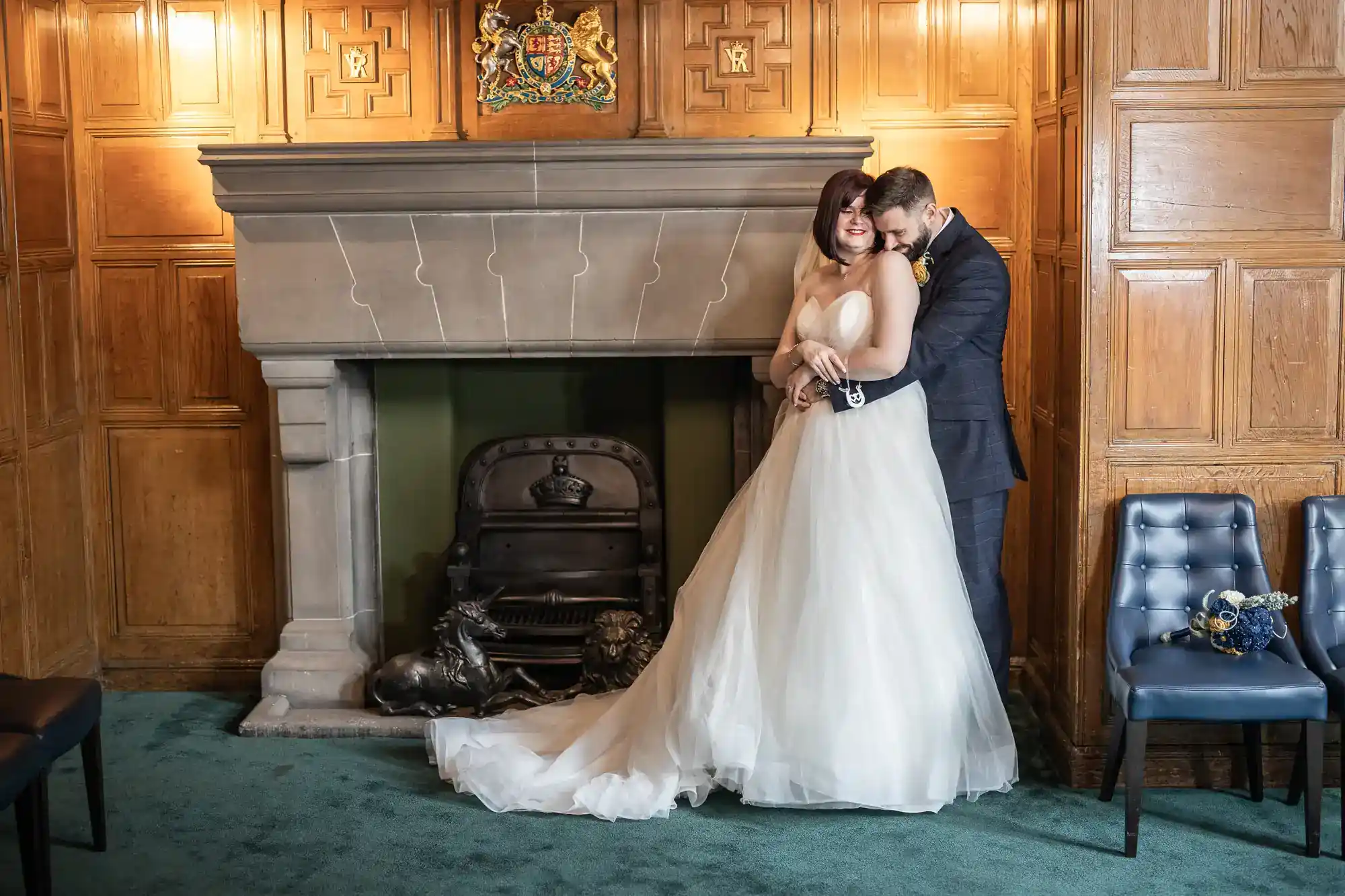 A bride in a white gown and a groom in a dark suit embrace and smile by a stone fireplace in a wood-panelled room. The bride’s train flows onto the green carpet. Two blue chairs and a bouquet are on the right. A crest hangs above the fireplace.
