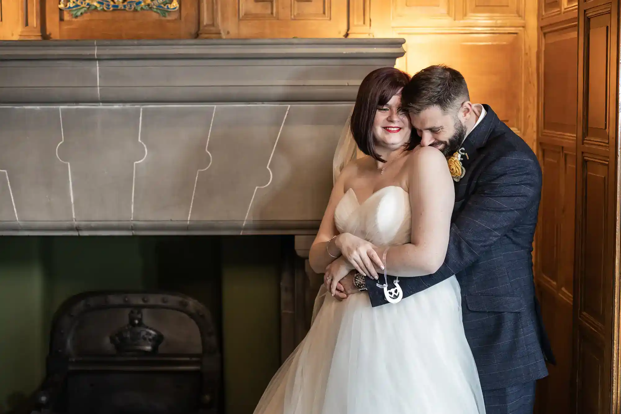 A bride in a strapless white gown and a groom in a dark suit embrace and smile in front of a carved stone fireplace with wood-panelled walls. The bride faces forward, whilst the groom hugs her from behind, both appearing joyful and relaxed.