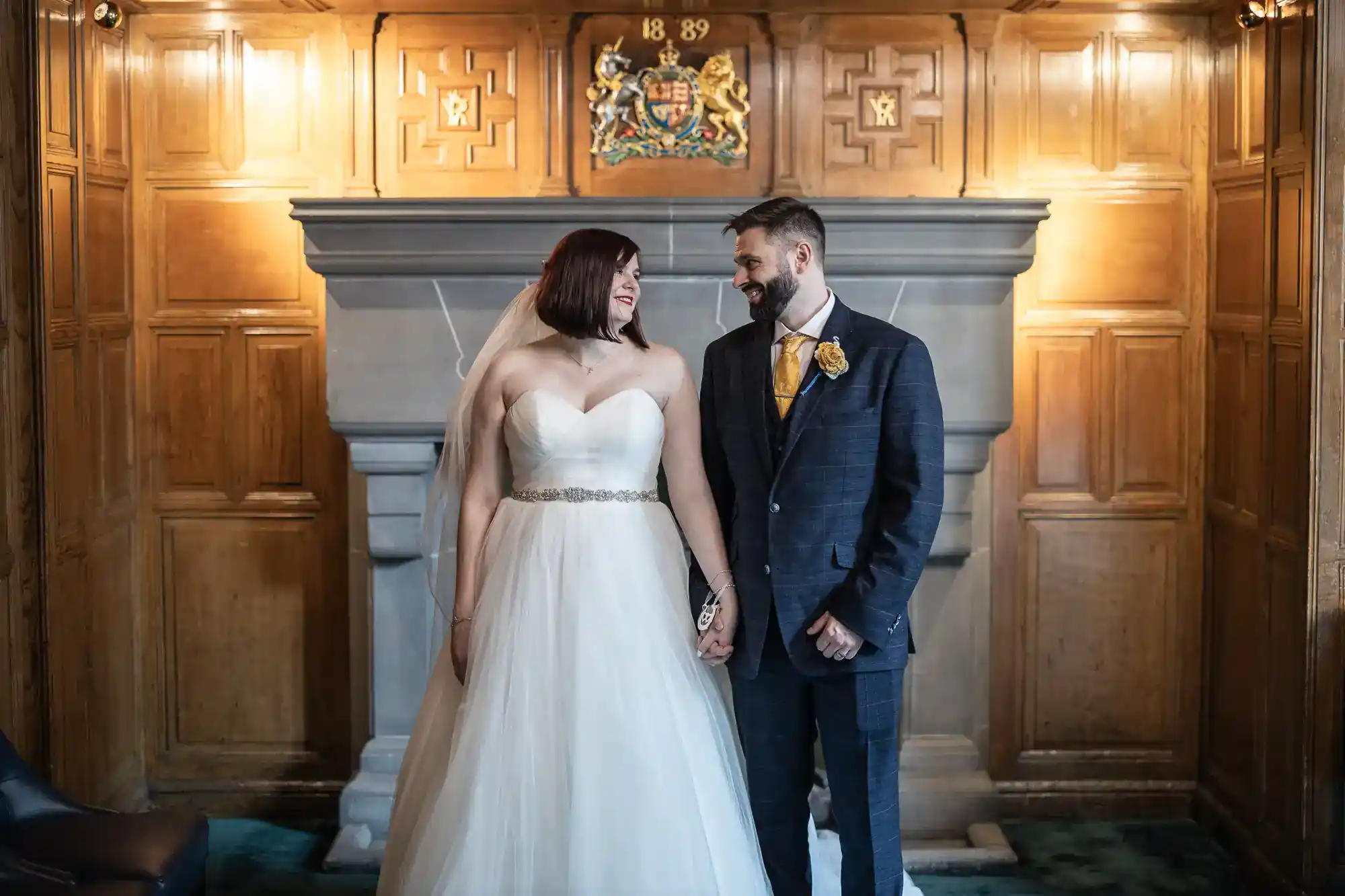 A bride in a strapless white gown and a groom in a dark checked suit stand holding hands, smiling at each other, in front of a fireplace with a crest and wood-panelled walls in a formal room.