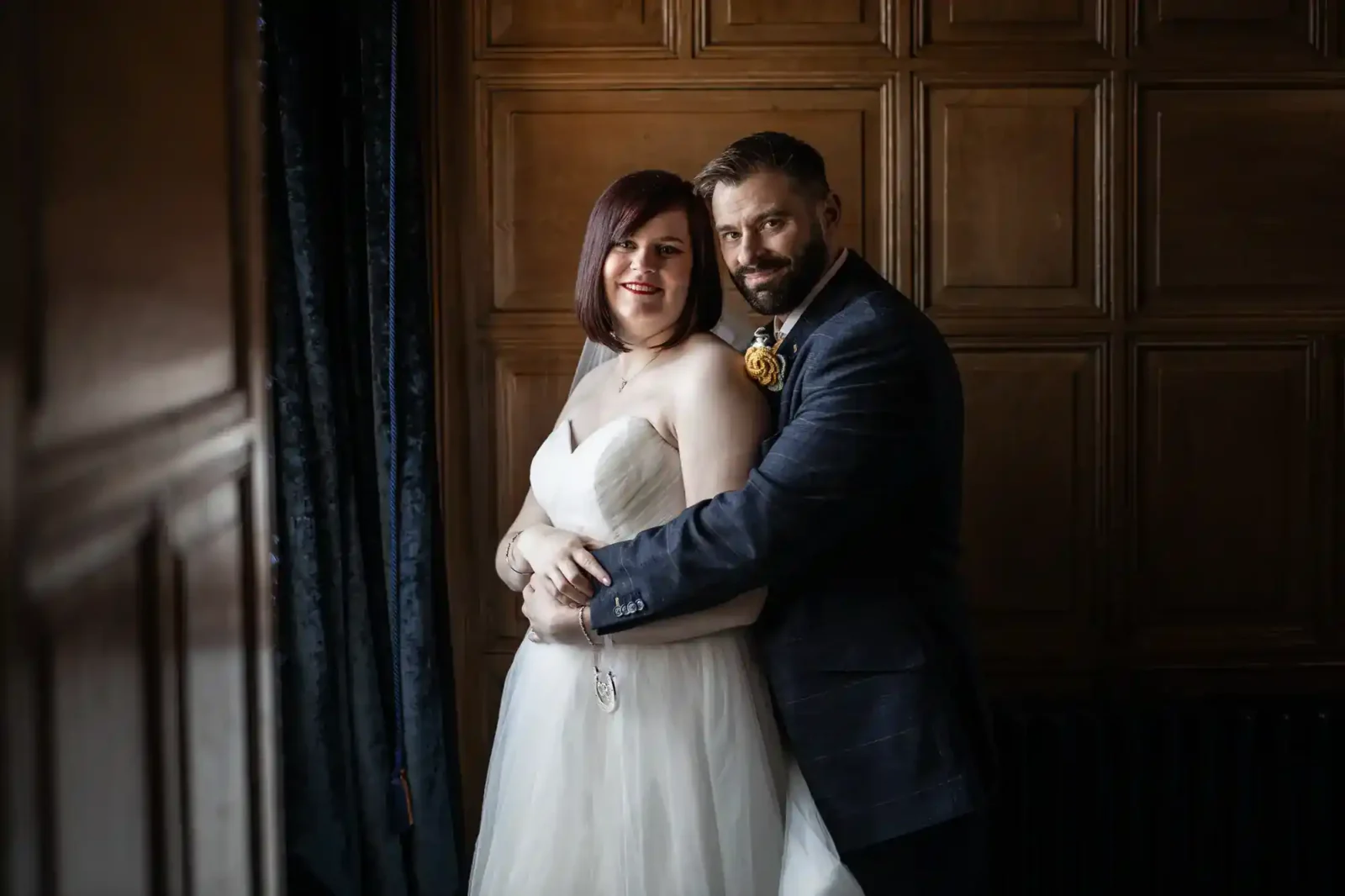 A bride in a white strapless dress and a groom in a dark suit embrace, smiling at the camera. They stand indoors in The Gatehouse Suite at Edinburgh Castle, by a dark, panelled wooden wall, with soft light illuminating them from the left.