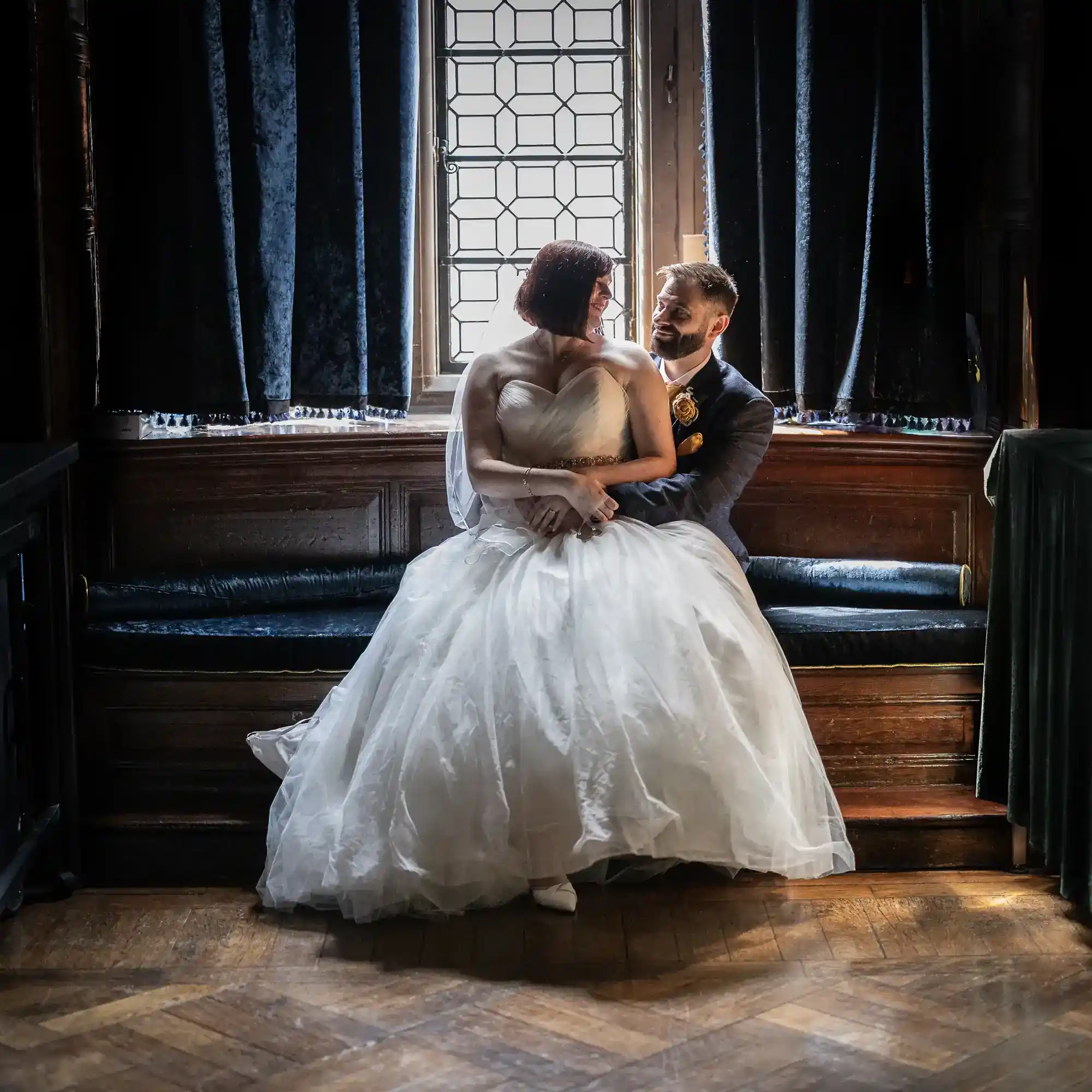 A bride in a white gown sits on a window seat beside a smiling groom in a suit. Soft daylight streams through the window behind them. They gaze lovingly at each other in a warmly lit, wood-panelled room at The Gatehouse, created by their Edinburgh Castle wedding photographer.