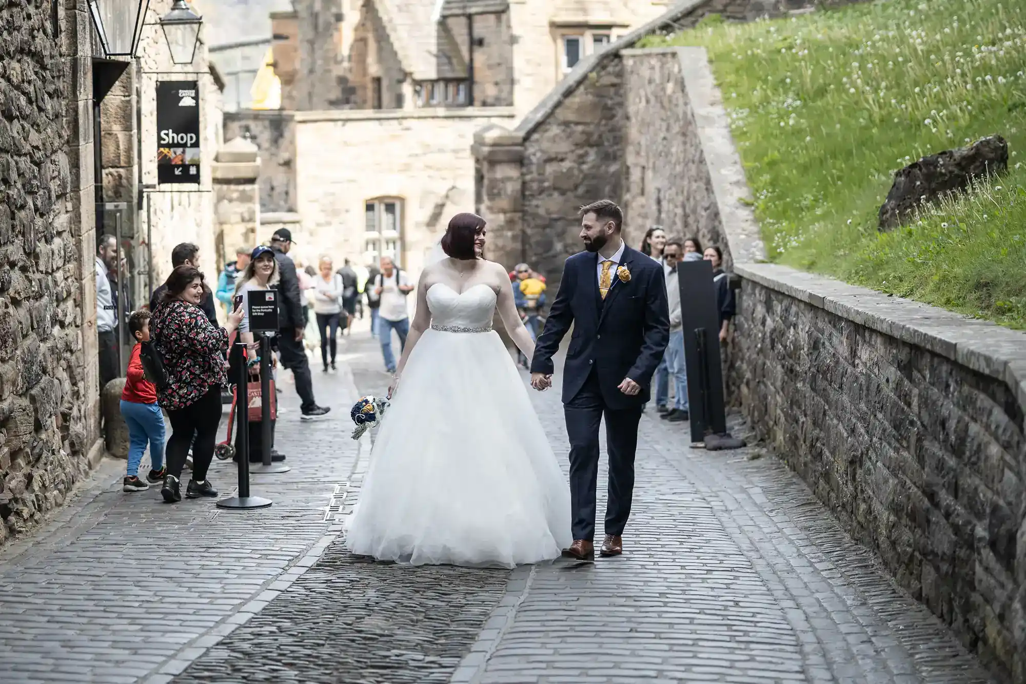A bride in a white dress and a groom in a dark suit walk hand in hand on a cobbled street, surrounded by stone walls. Onlookers stand nearby, some taking photos, as the couple smile at each other. The scene has a historic, festive atmosphere.