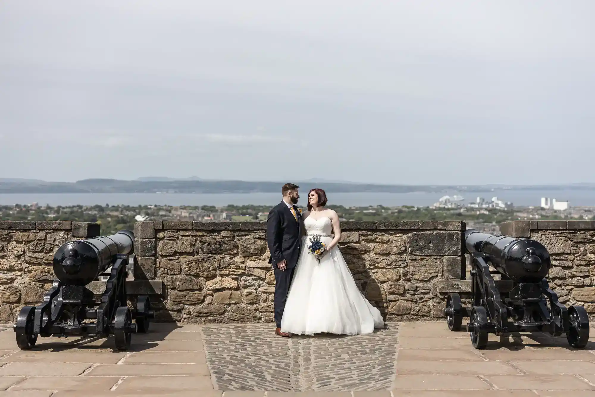 A bride in a white dress and groom in a dark suit stand together on a stone patio between two old cannons, with a stone wall behind them. City buildings and water are visible in the distant background under a pale sky.