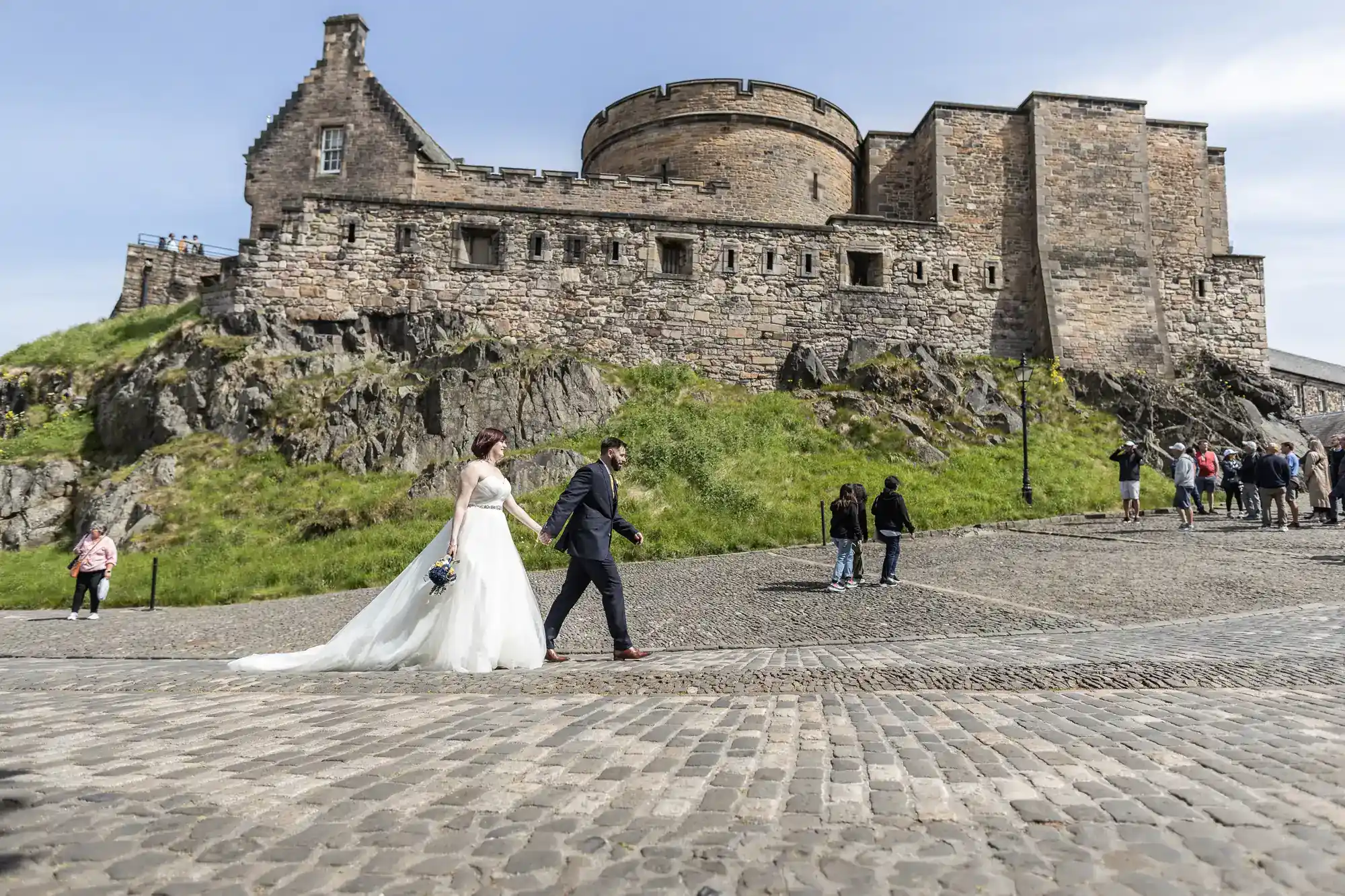 A bride and groom walk hand-in-hand in front of a historic stone castle on a sunny day. The bride wears a white gown; the groom has a suit. Tourists and visitors walk nearby. Green grass and cobblestone pavement are visible in the scene.