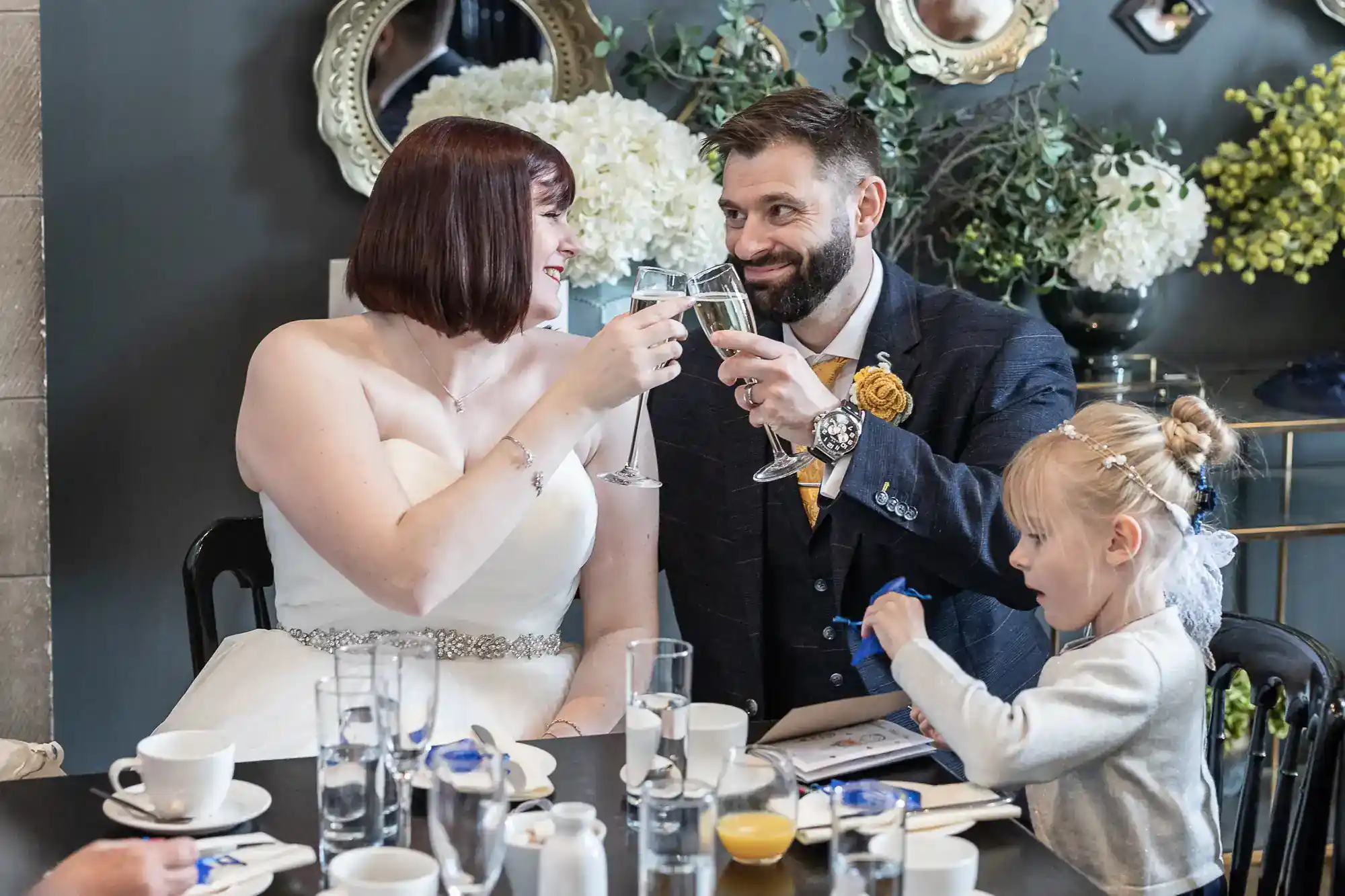 A bride and groom clink glasses and smile at each other at a wedding reception table, whilst a young girl with blonde hair sits beside them, drawing. Elegant decor, flowers, and mirrors are visible in the background.