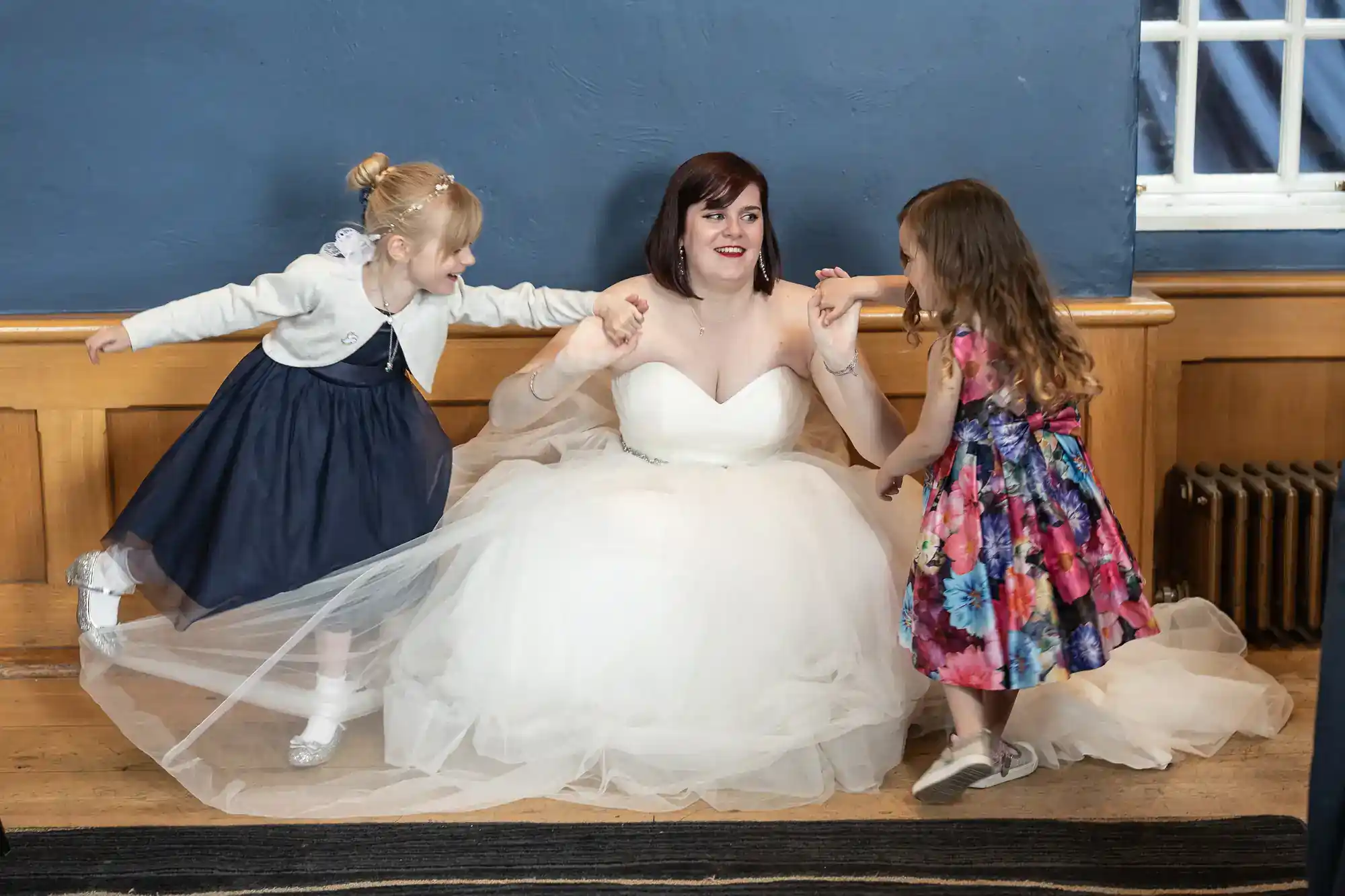 A smiling bride in a white gown sits on the floor against a blue wall, holding hands with two playful young girls—one in a navy dress and cardigan, the other in a colourful floral dress. All appear joyful and engaged in fun together.