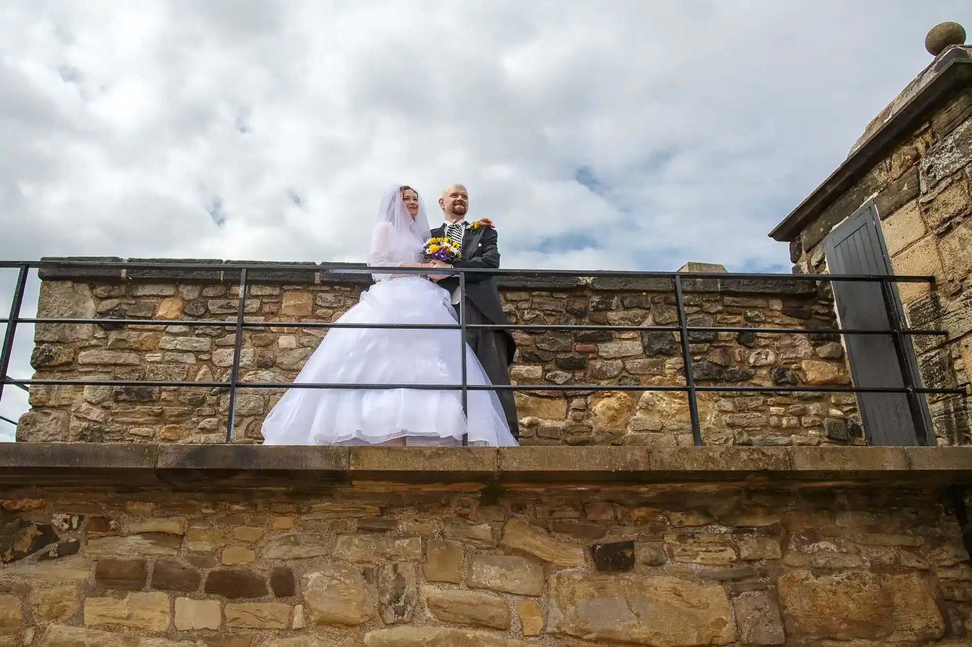 A bride and groom stand on a stone balcony in wedding attire, with the bride in a white dress holding a bouquet. They are smiling, surrounded by a stone wall and cloudy sky above. The scene is outdoors, evoking a joyful, celebratory atmosphere.