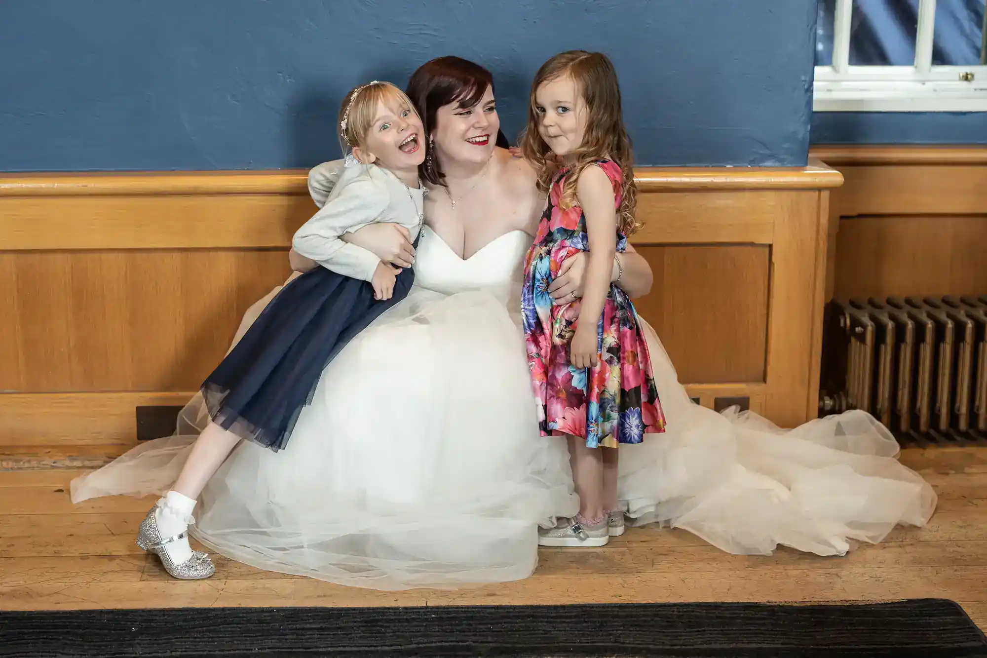 A bride in a white dress sits smiling on the floor, hugging two young girls. One girl wears a navy dress with a white jacket, the other a colourful floral dress. They pose closely together in front of a wooden wall with a blue backdrop and a window.