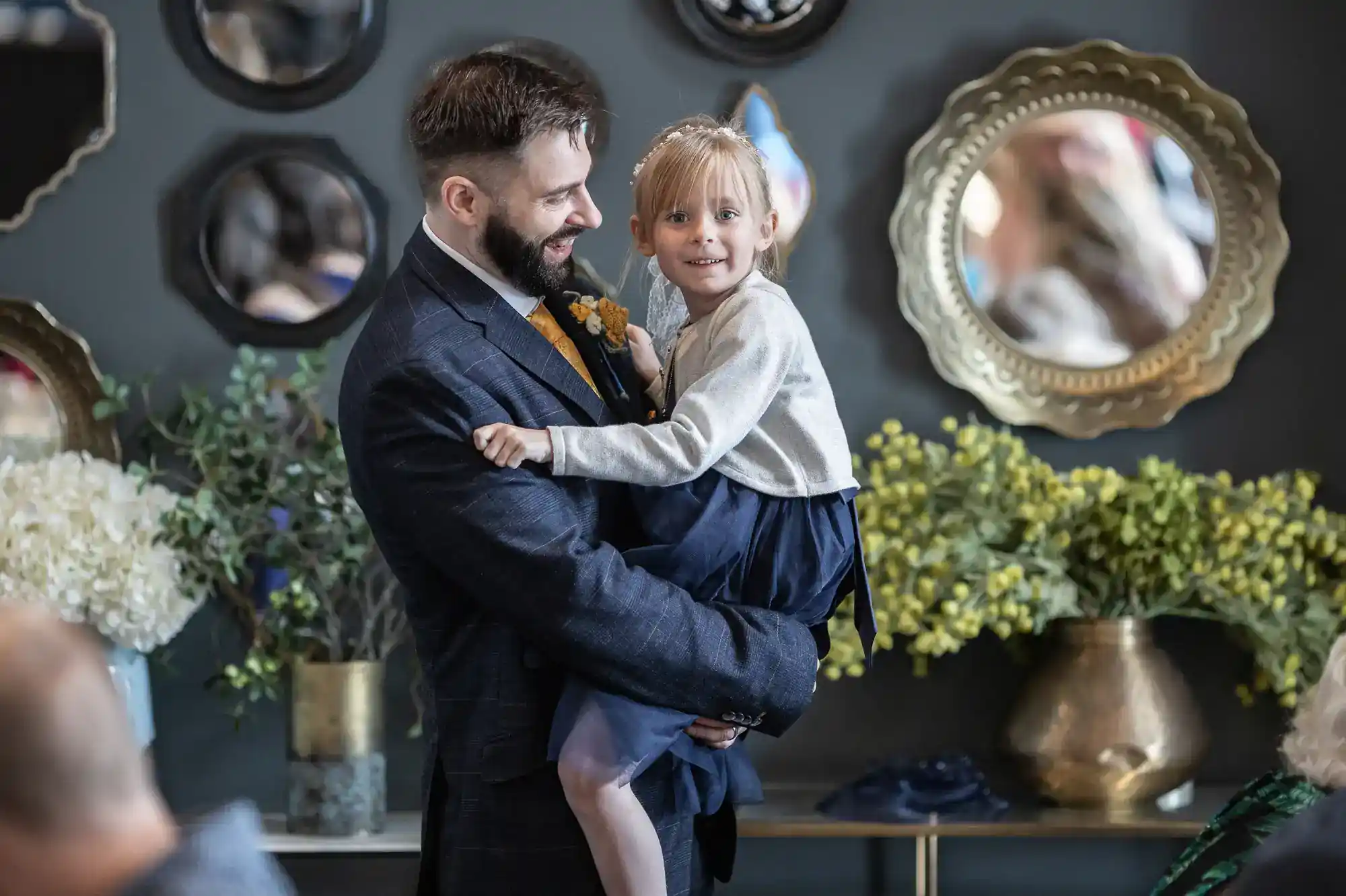 A man in a suit holds a smiling young girl in a blue dress. They stand indoors, surrounded by round mirrors on a grey wall and flower arrangements in gold vases. The mood is joyful and warm.