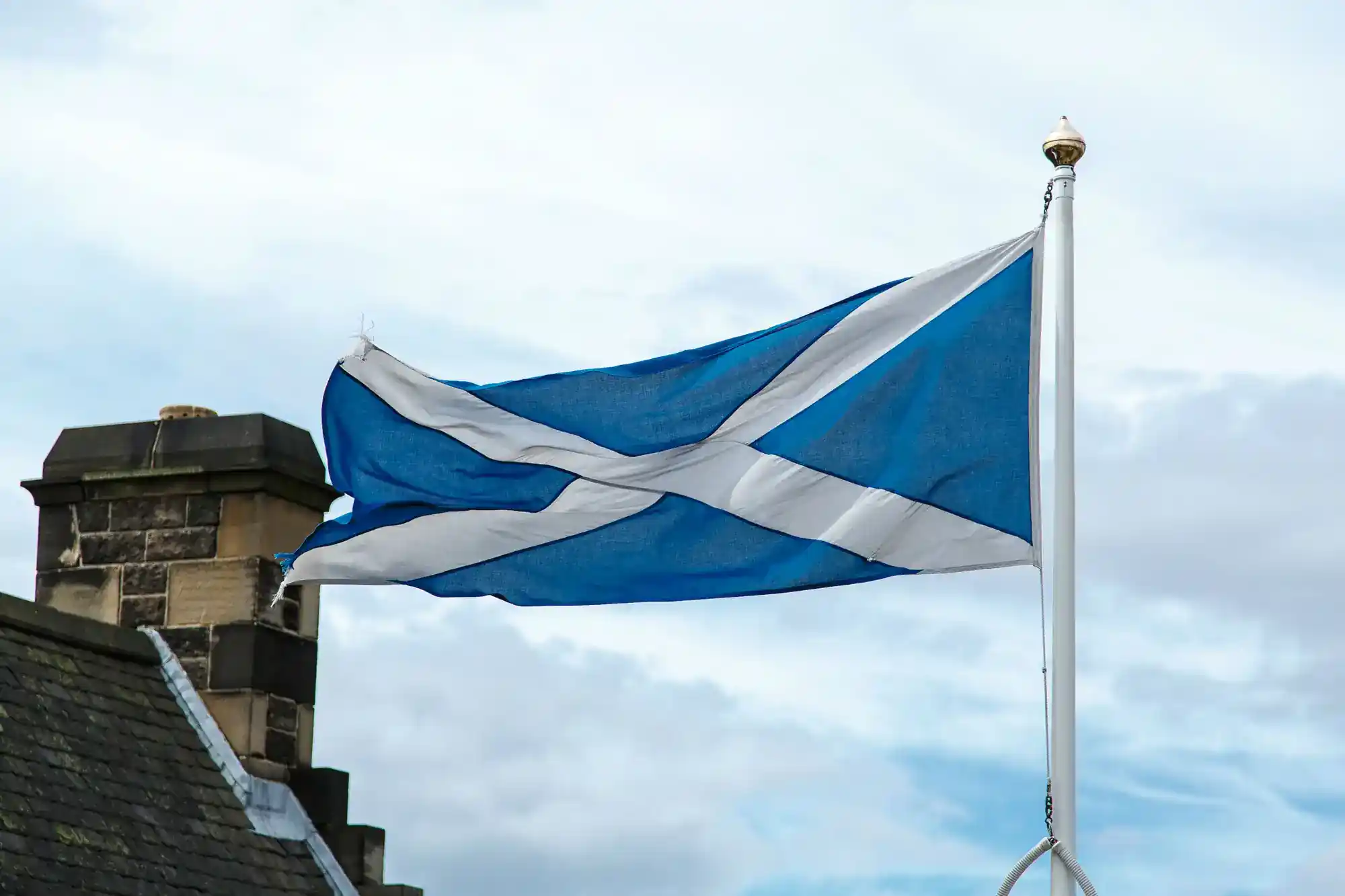 The Scottish flag, a blue field with a white diagonal cross, flies on a flagpole against a cloudy sky. Part of a stone building with a chimney is visible on the left side of the image.