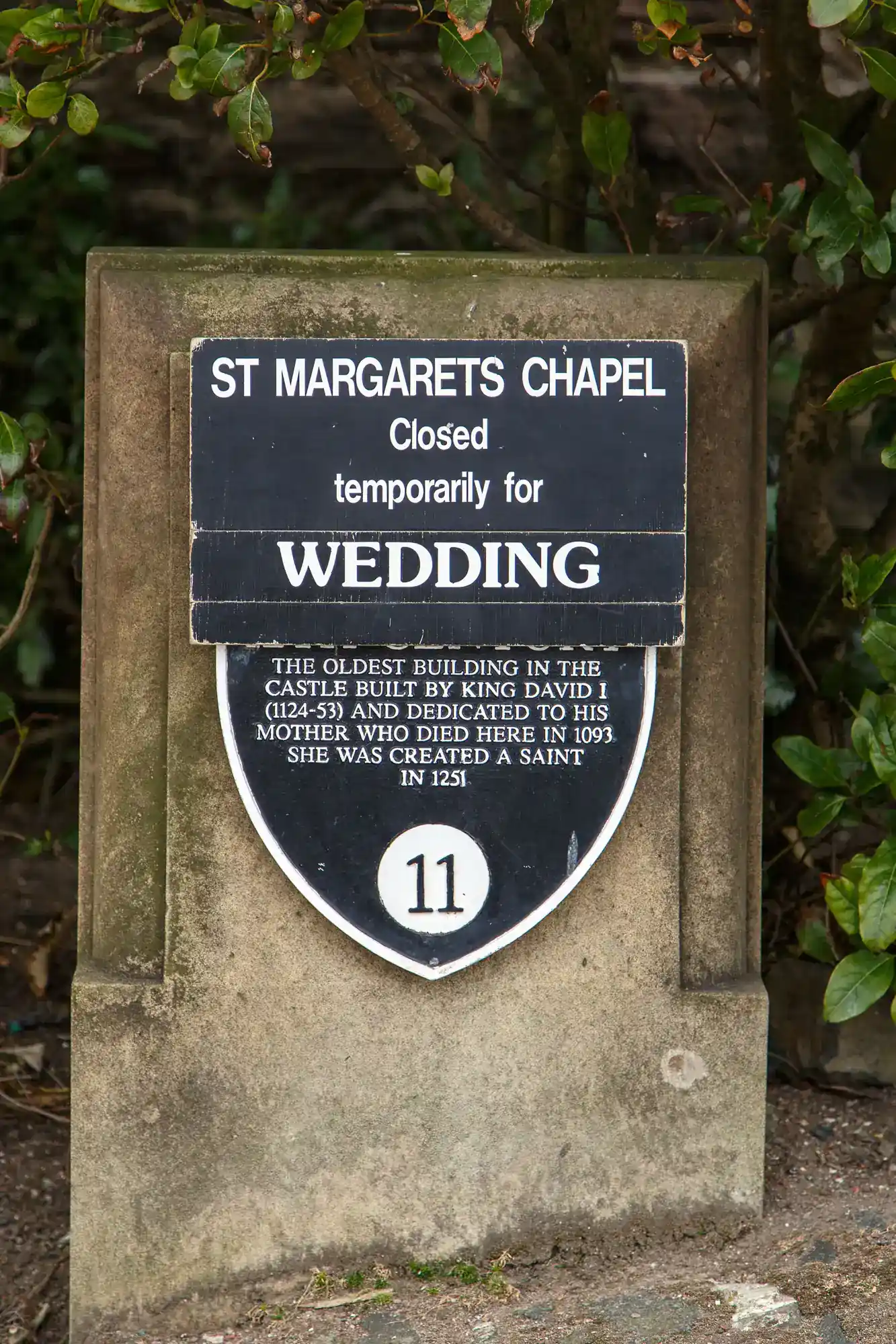 A sign at St Margaret’s Chapel reads: “Closed temporarily for wedding.” Below, a plaque notes it’s the oldest building in the castle, built by King David I and dedicated to his mother, with “11” displayed at the bottom. Lush greenery frames the sign.