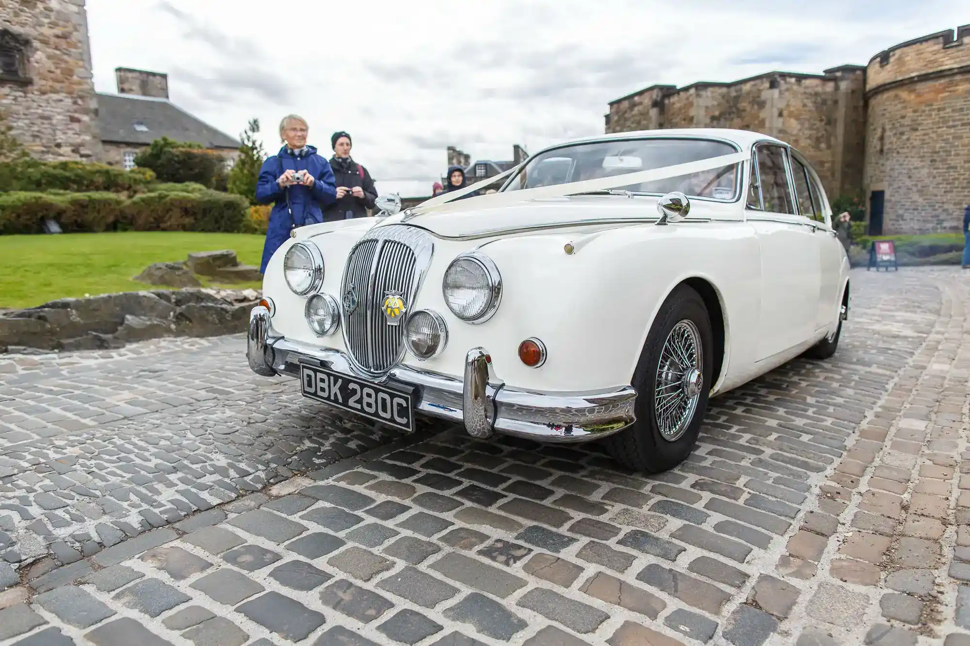 A white vintage Jaguar Mark 2 car with registration plate DBK 280C is parked on cobblestones near a stone building. Three people stand in the background, two taking photos. The sky is overcast and greenery is visible on the left.