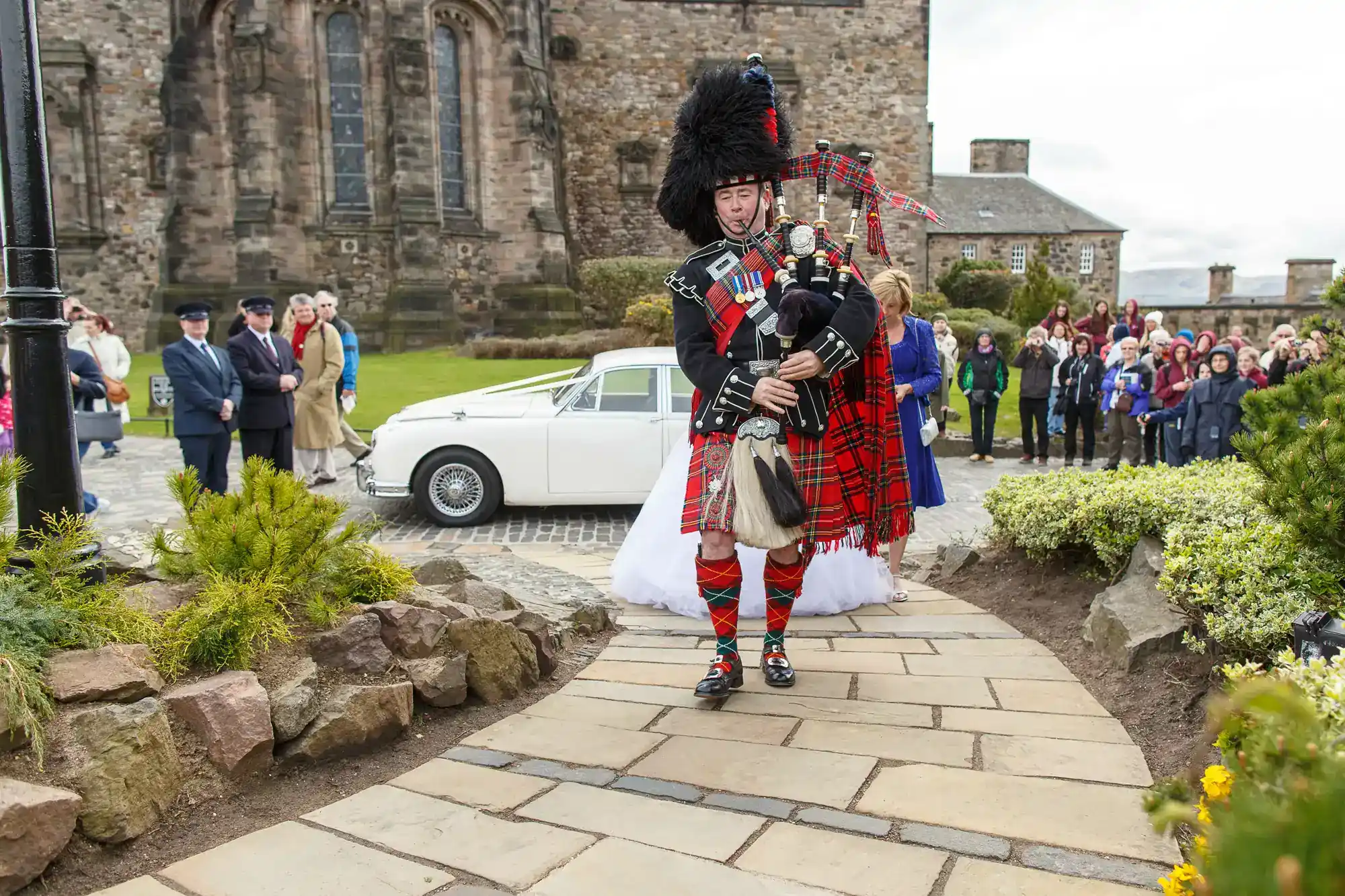 A bagpiper in traditional Scottish attire leads a bride in a white dress along a stone path. Behind them, guests stand near a white vintage car parked by an old stone building. The setting is outdoors, and the sky appears cloudy.