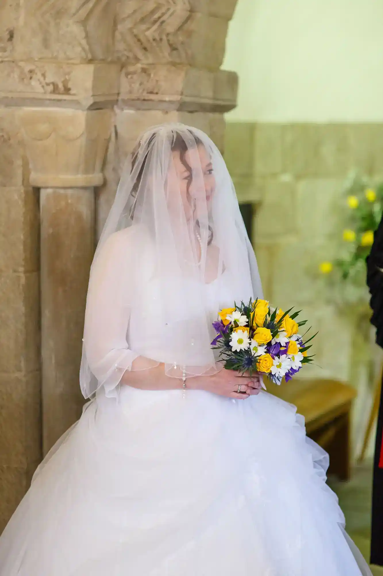 A bride in a white dress and veil stands indoors, holding a bouquet of yellow, purple, and white flowers. The background features stone walls and pillars, with soft lighting. She appears to be smiling and is partially turned towards someone out of frame.