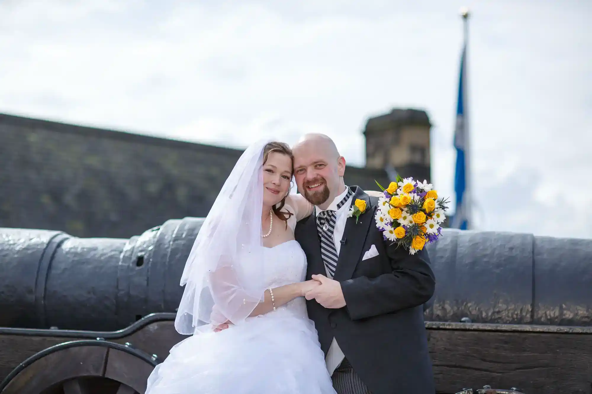 A bride in a white dress and veil and a groom in a black suit and striped tie stand smiling together, holding hands and a bouquet of yellow and white flowers, in front of a large black cannon and a building with a flag.
