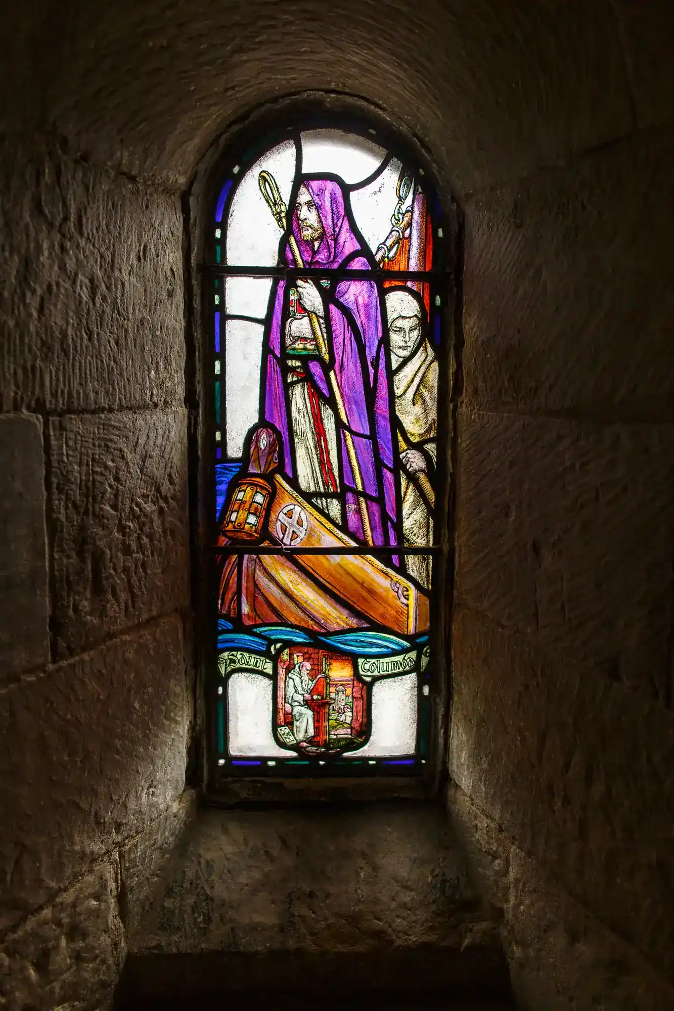 A stained glass window set in a stone wall depicts a robed figure in purple holding a staff, standing beside a kneeling figure. The scene includes a shield and coat of arms at the bottom, surrounded by intricate coloured glass details.
