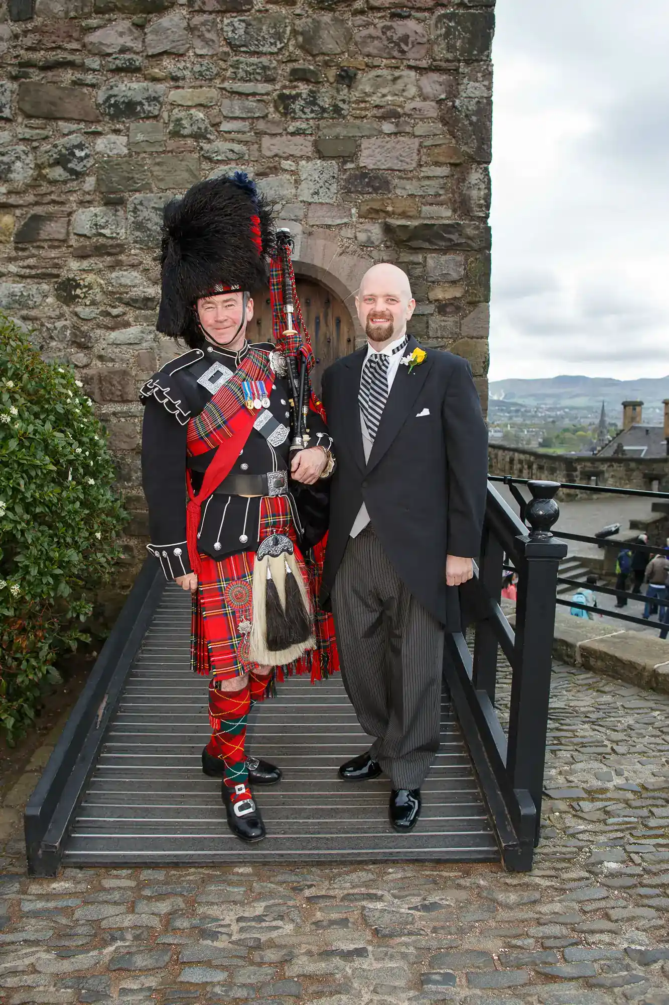 A man in a black dinner jacket stands beside a bagpiper in full traditional Scottish attire, including a feathered hat, red tartan kilt, and sporran, on a ramp by a stone building. Both are smiling and posing for the photo outdoors.