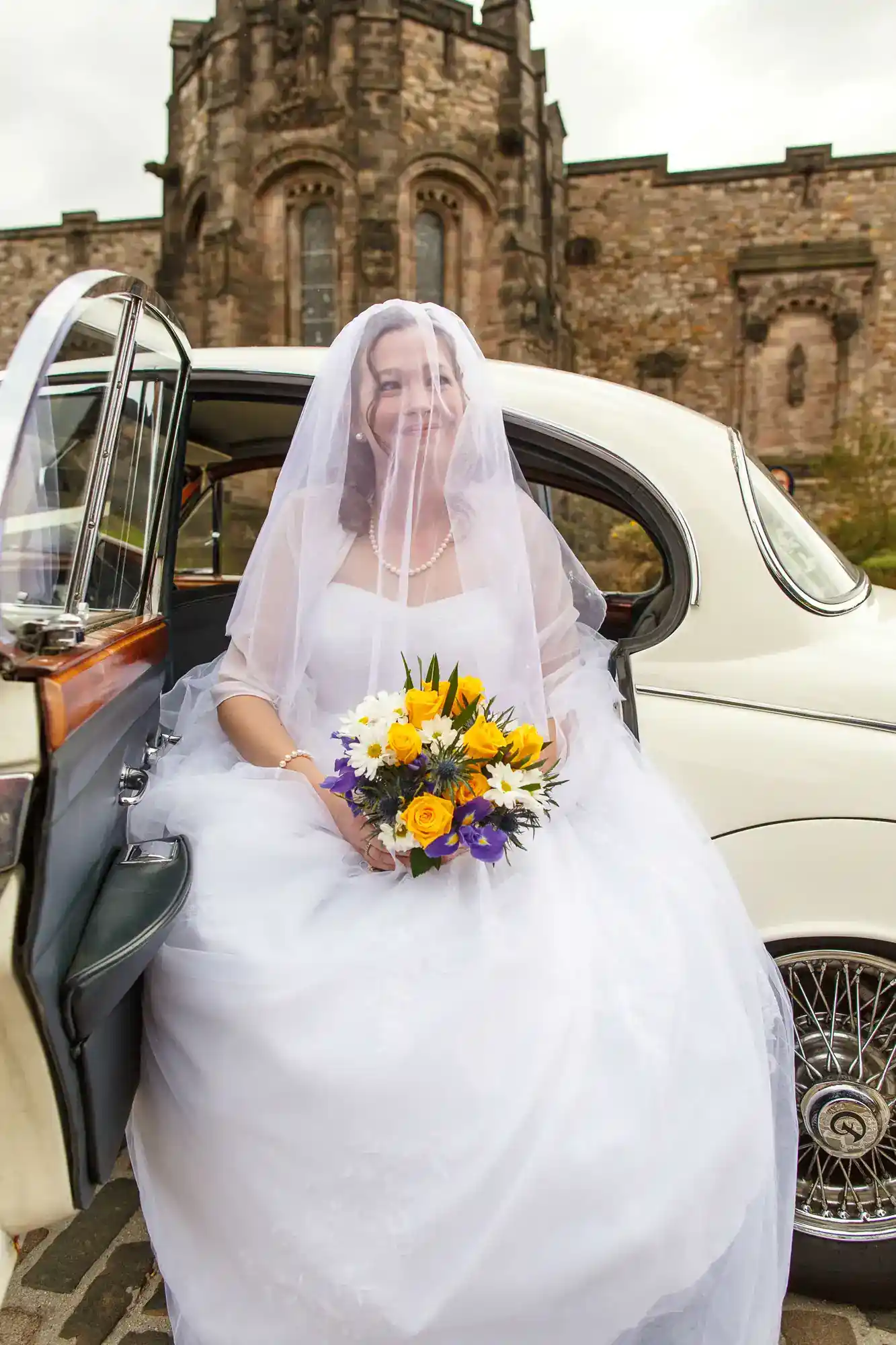 A bride in a white wedding dress and veil sits in a vintage car’s open door, smiling and holding a bouquet of yellow, white, and purple flowers. A historic stone building with arched windows is visible in the background.