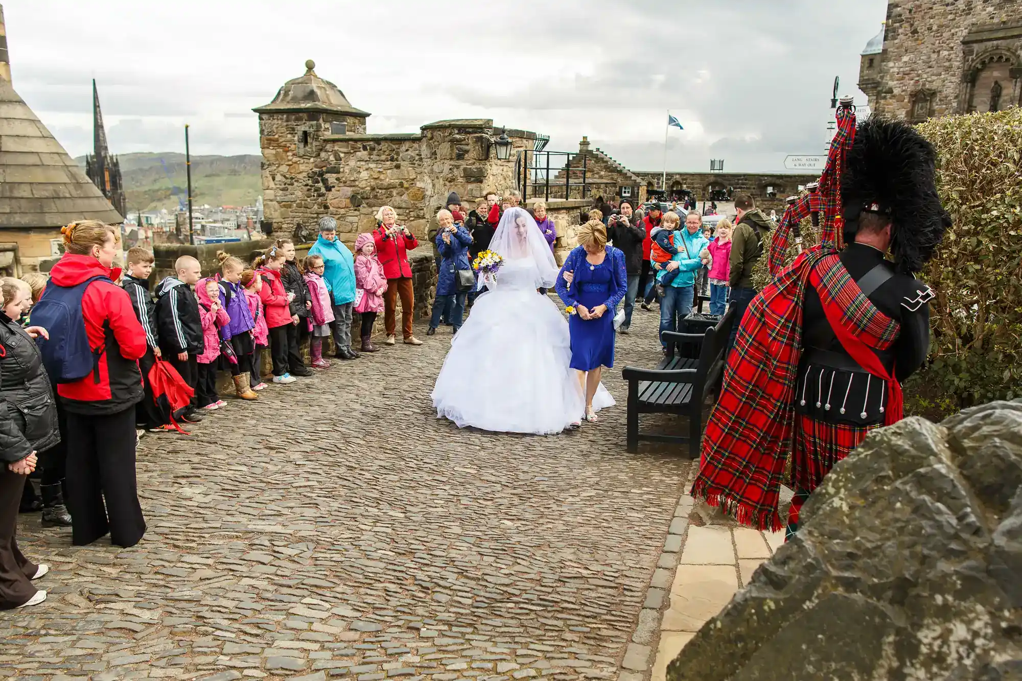 A bride in a white dress walks with a woman in blue on a cobbled path, surrounded by onlookers. Children and adults watch; a bagpiper in traditional Scottish dress plays nearby. The scene is outdoors near historic stone buildings.