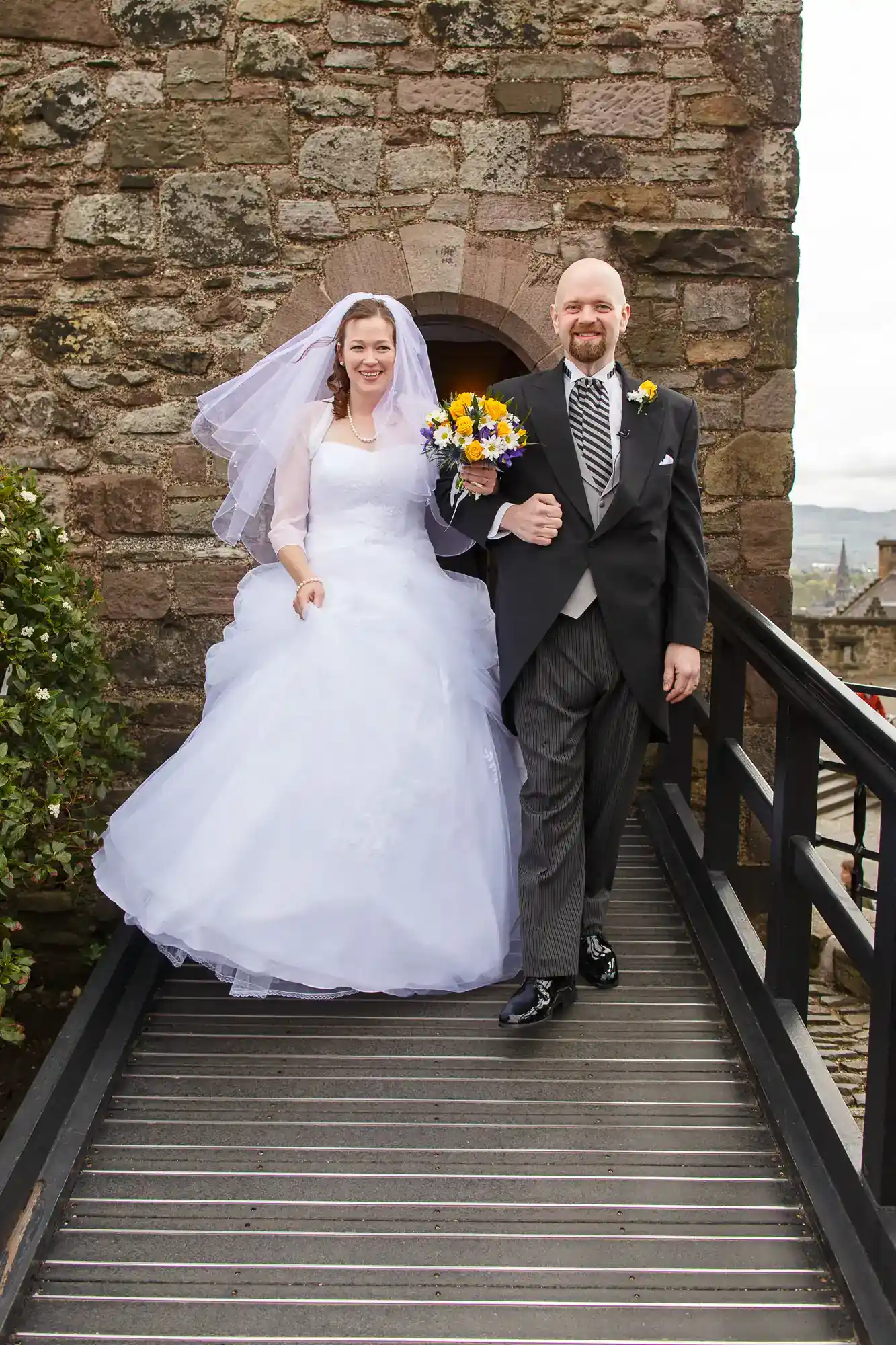A bride in a white gown and veil stands arm-in-arm with a man in a black suit, striped trousers, and a tie. He holds a bouquet of yellow and purple flowers. They smile, standing on a bridge with a stone wall in the background.