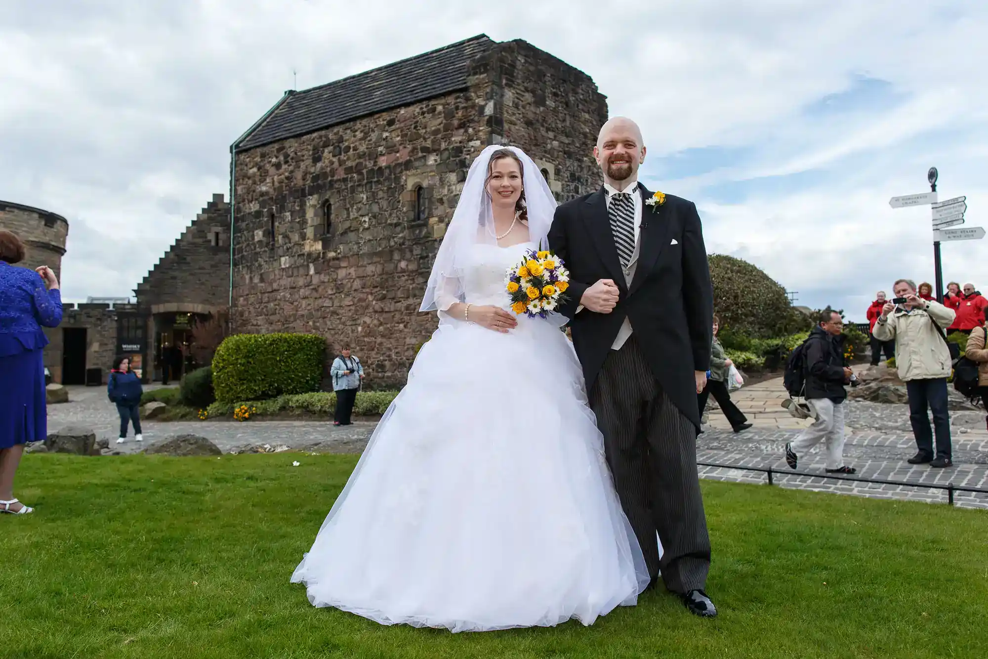A bride in a white gown and veil holds yellow and blue flowers, standing arm-in-arm with a groom in a black jacket, striped trousers, and yellow buttonhole outside a stone building, with people and a cloudy sky in the background.