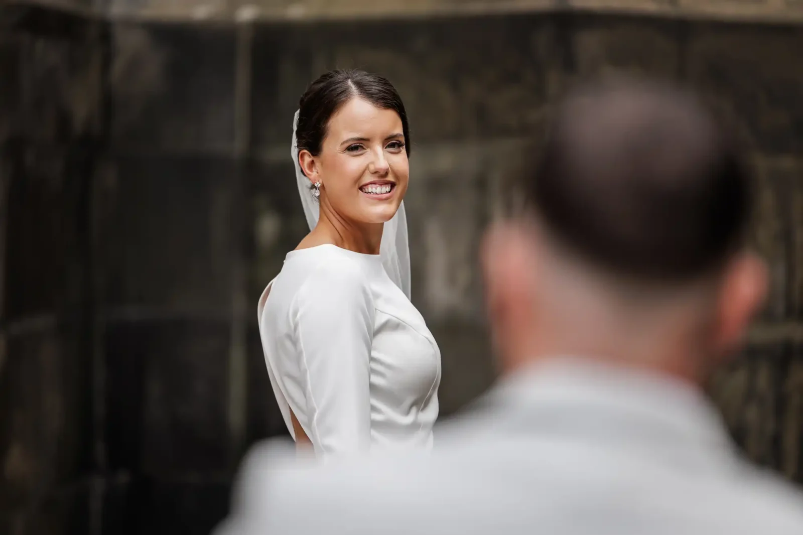 A smiling bride in a white dress and veil looks over her shoulder towards the camera. Her dark hair is styled up, and she wears pearl earrings. A person with short hair, seen from behind and out of focus, stands in the foreground. The background is stone.